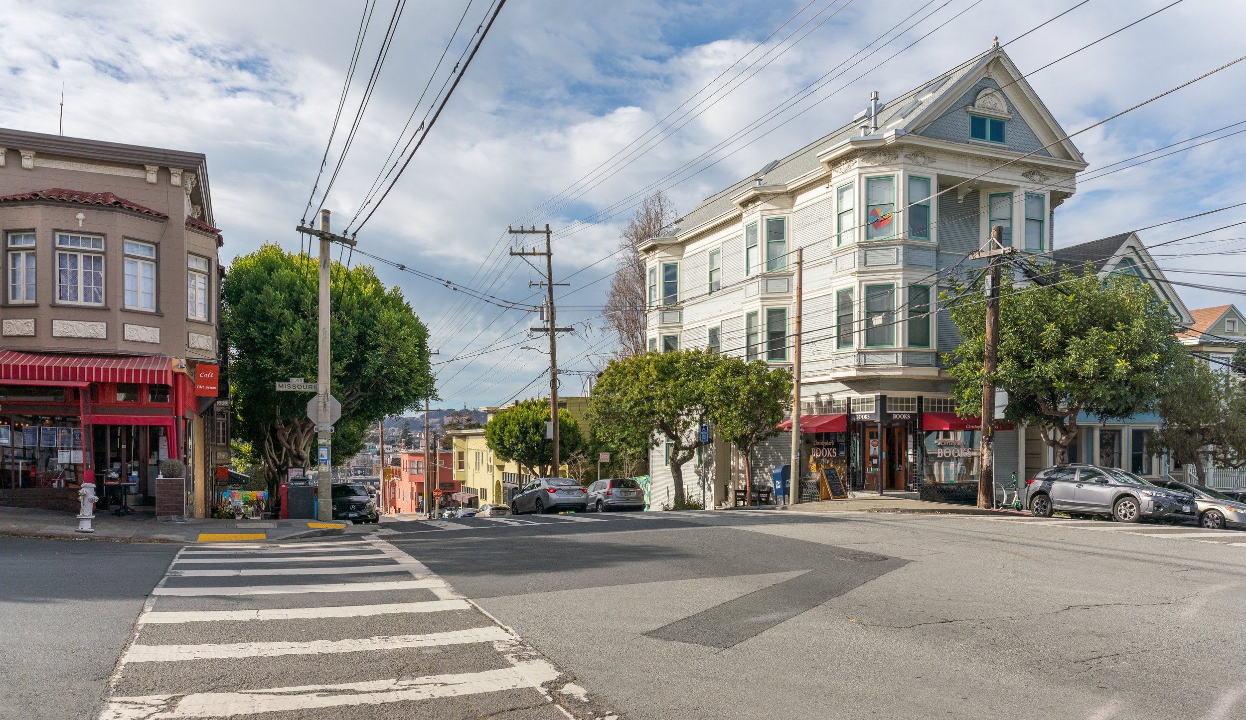 Street view of a neighborhood with shops, trees, parked cars, and houses under a partly cloudy sky.