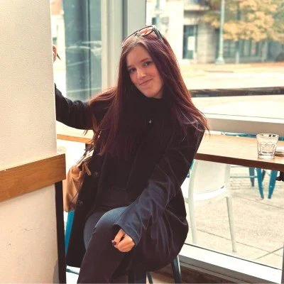 A young woman with long brown hair sitting indoors by a large window, smiling at the camera, wearing a black jacket and glasses, with a table and a glass behind her.