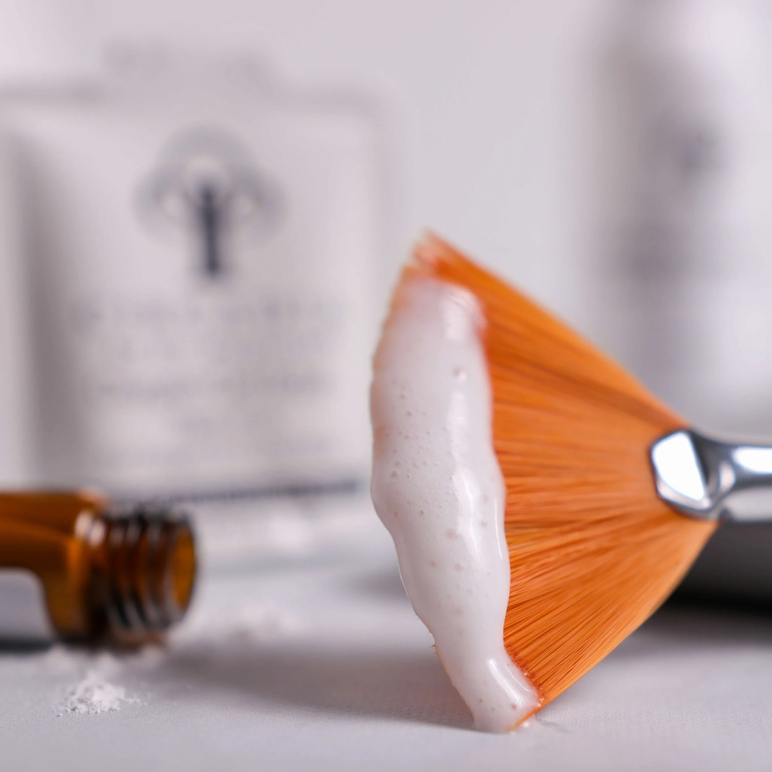 A shaving brush with orange bristles and soap foam on it, placed on a white surface with bottles of shaving cream or lotion in the background.