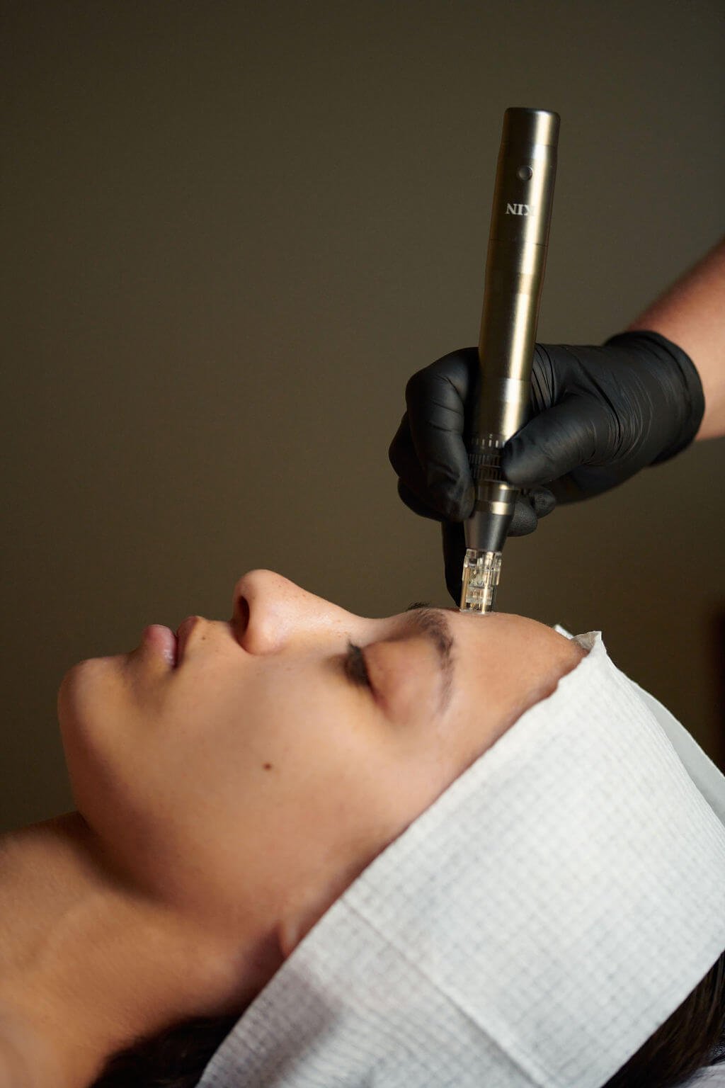 A person receiving a facial treatment with a handheld device on their forehead, lying down with eyes closed, wearing a white headband, and a person in black gloves operating the device.