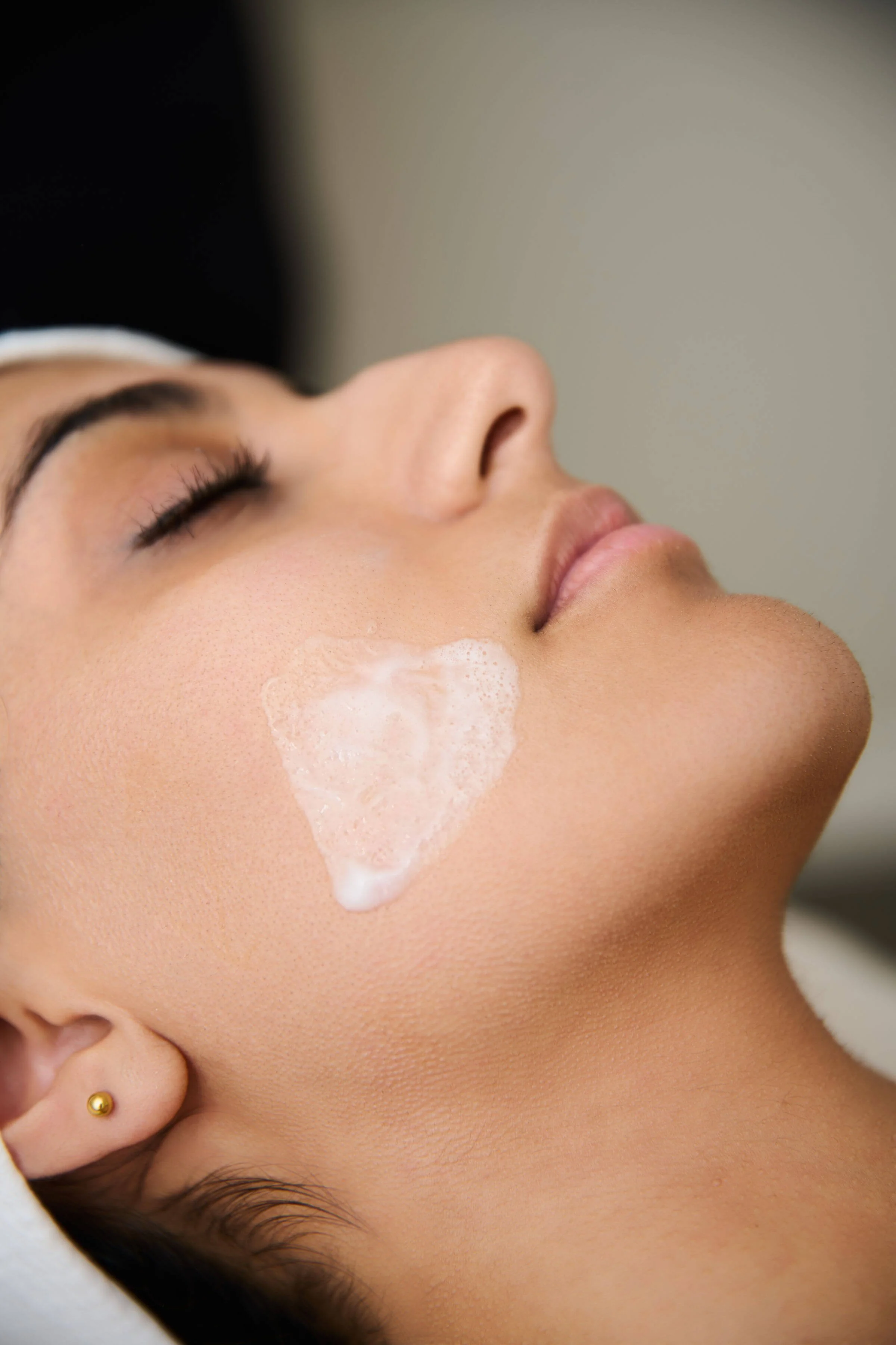 Close-up of a woman with her eyes closed, lying back, having a facial treatment with foamy cleanser on her cheek.