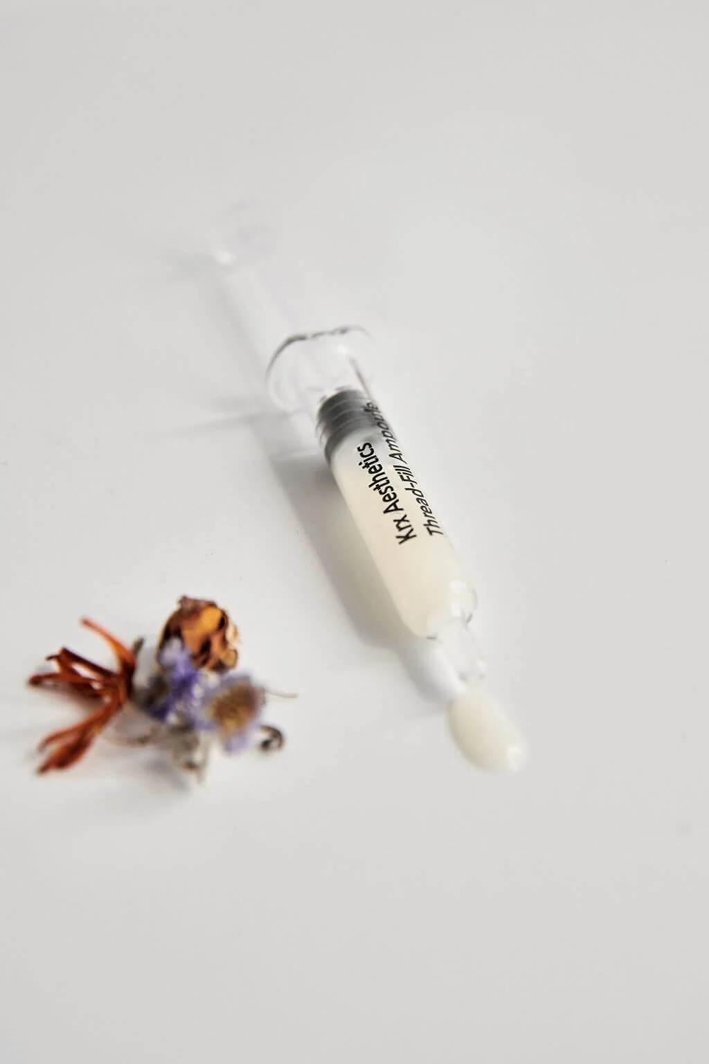 A syringe filled with a white substance placed on a white surface next to dried flowers.