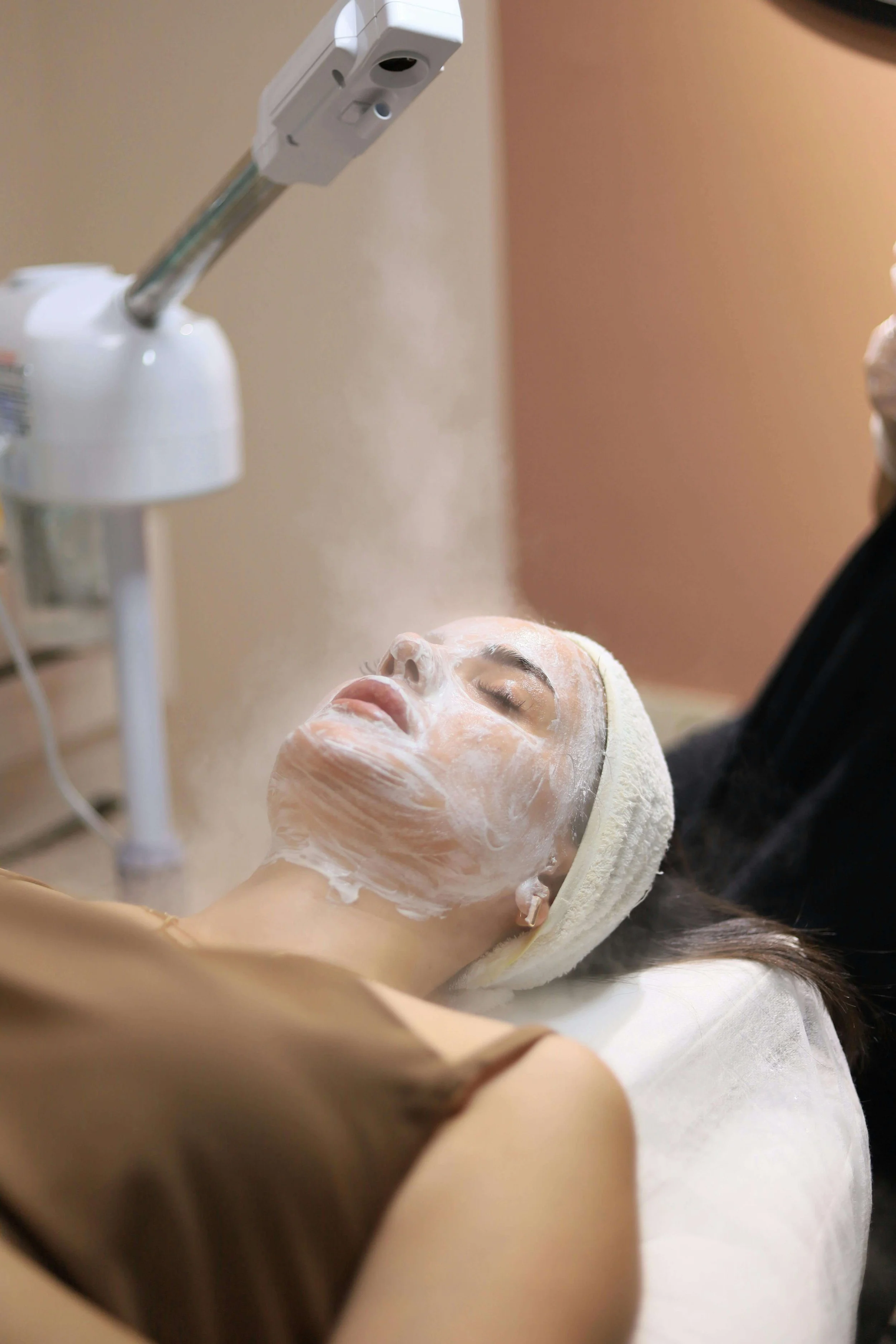A woman receives a facial treatment with steam or mist being applied to her face at a spa or skincare clinic.