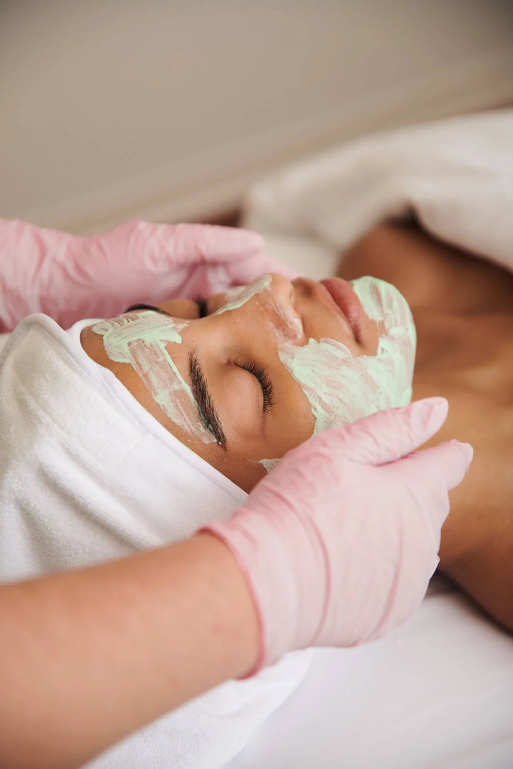 A woman receives a facial treatment with a white facial mask applied to her face, while a skincare professional wearing pink gloves gently handles her head.