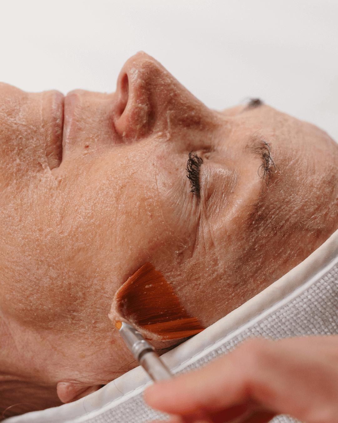 A person receiving a facial treatment with a brush on their cheek, lying down with their eyes closed.
