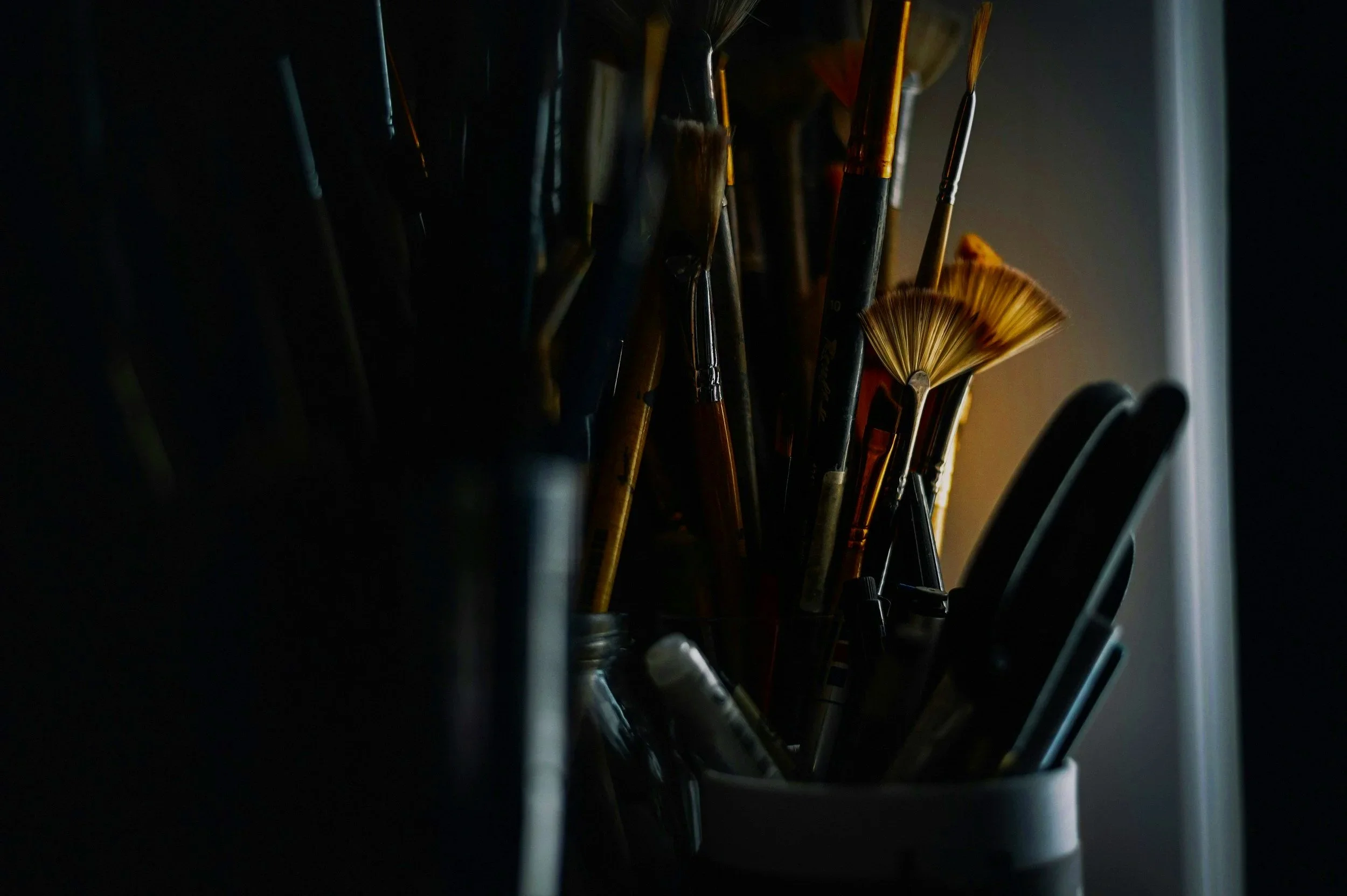A collection of paintbrushes in a container, with some brushes illuminated by a soft warm light and many in shadow, creating a contrast between light and dark.