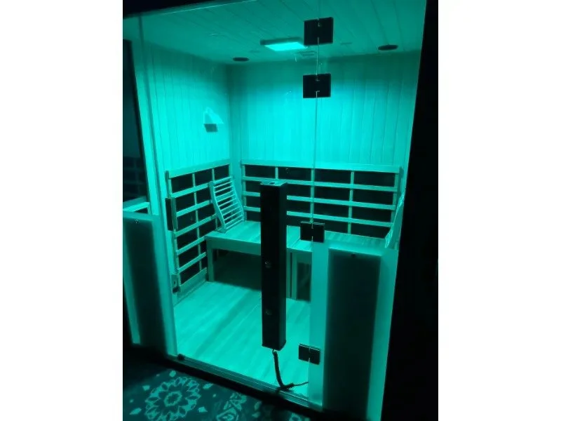 halotherapy infrared sauna in a sauna room illuminated with blue light, featuring wooden benches along three walls, a glass door with black hinges and handle, and a floor with a patterned rug outside the enclosure.