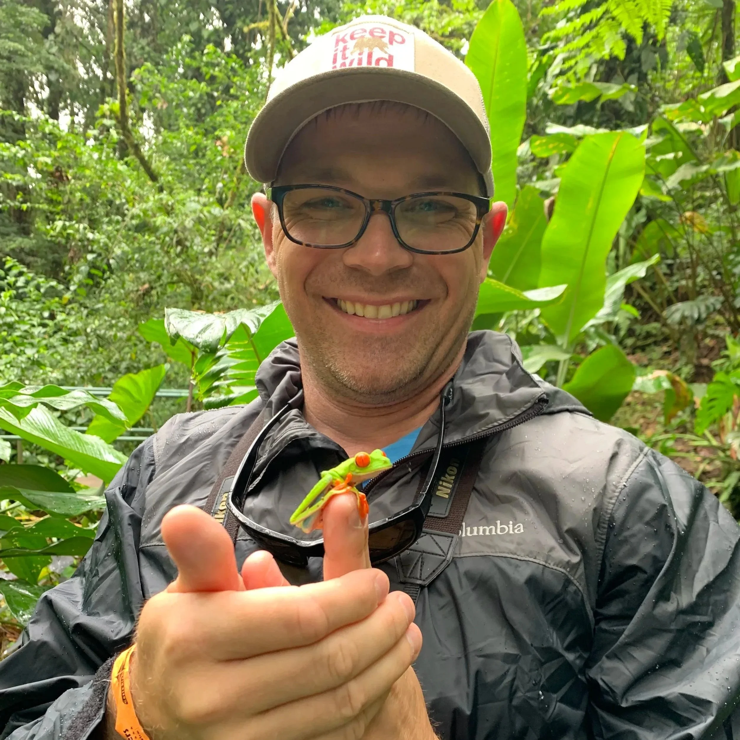A smiling man wearing glasses and a beige baseball cap with a red logo, standing in a lush green jungle, holding a colorful lizard in his hand.
