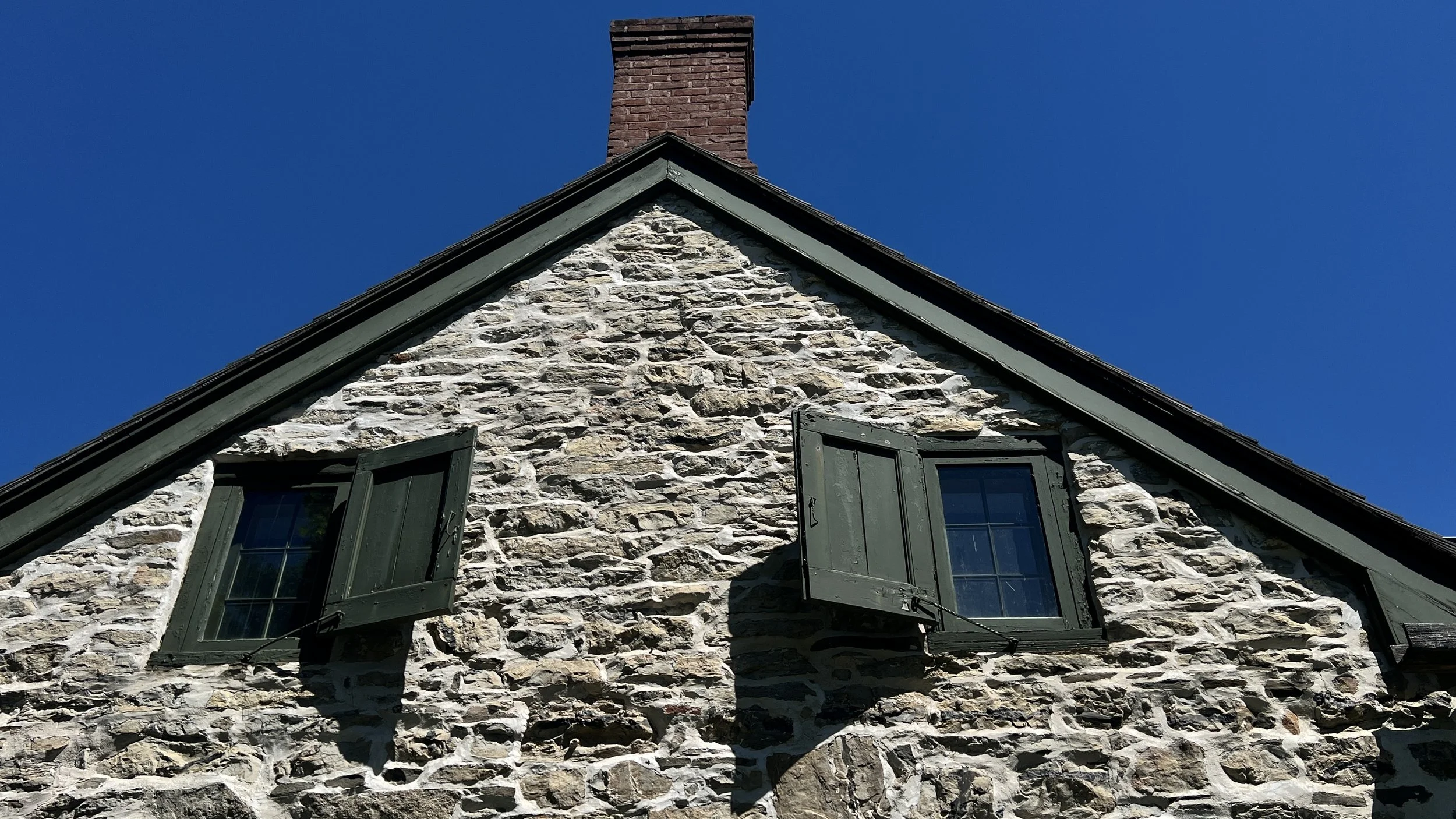 Close-up of a stone house with green window shutters, sloped roof, and a brick chimney against a clear blue sky.