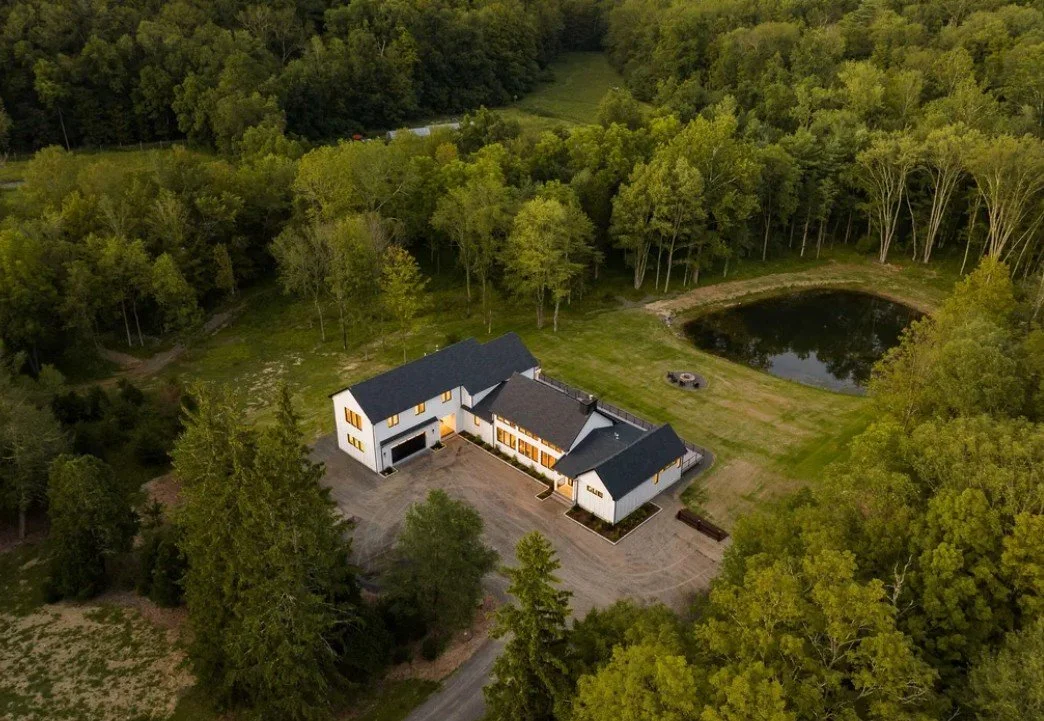 A large, modern white house with black roof nestled in a green forested area. The house features multiple sections, large windows with yellow curtains, and a gravel driveway. Nearby, there is a small pond and an open grassy space with a fire pit. Tall trees surround the property, creating a secluded setting.