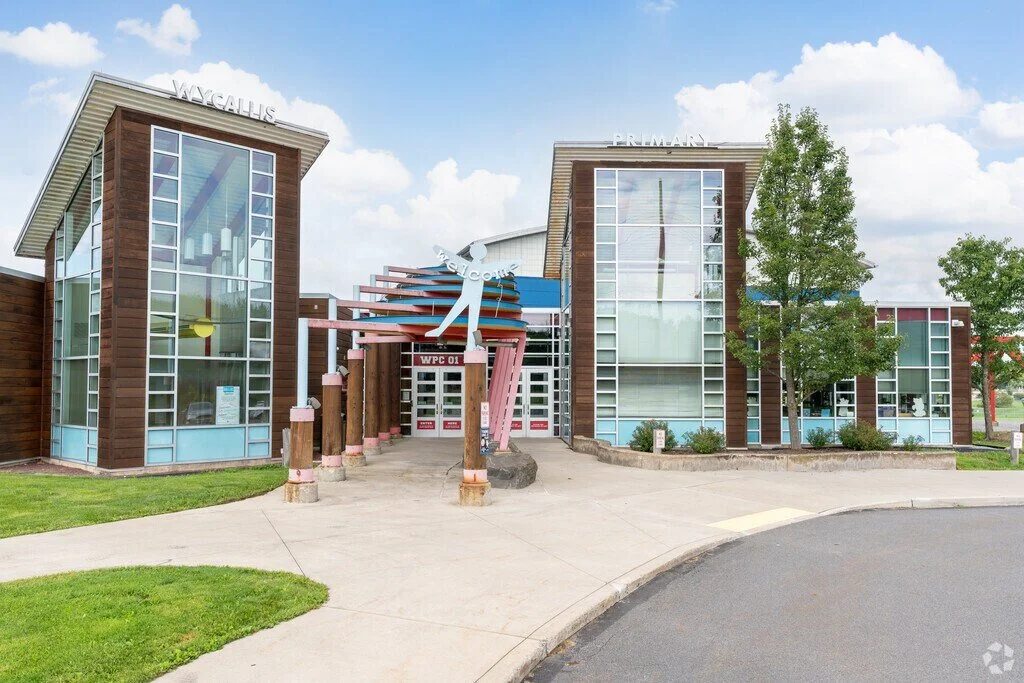 Modern school building with large glass windows, a colorful welcoming sculpture, and trees in front, labeled 'Wycallis' and 'Primary'