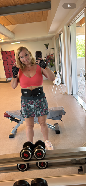 Frau macht Selfie im Fitnessstudio vor Spiegel, trägt Sportkleidung, trainer, mit Kurzhanteln im Vordergrund, Fenster mit draußen sichtbarem Balkon im Hintergrund.