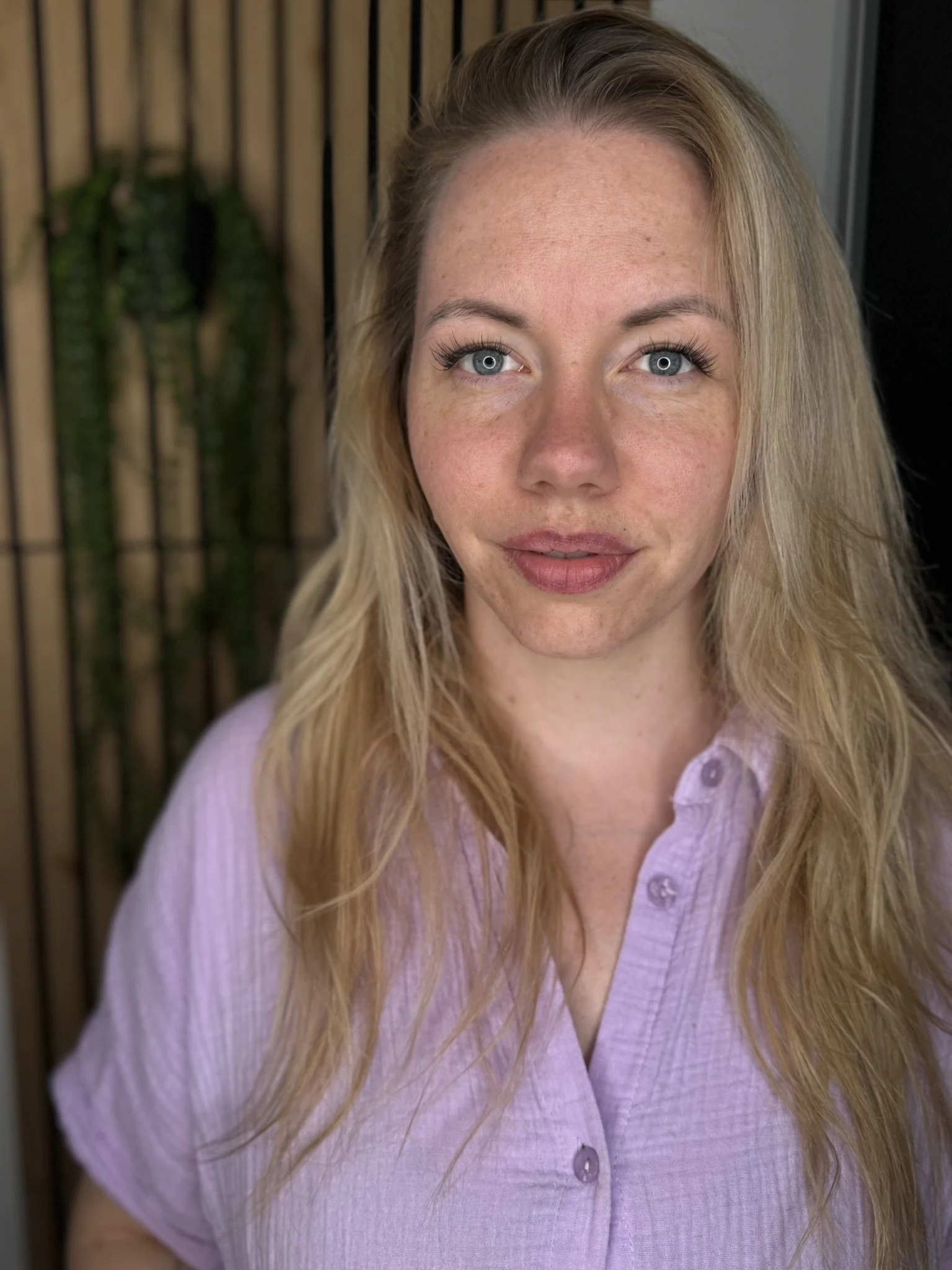 Nahaufnahme eines Mädchens mit hellblauen Augen, blonden Haaren und einem rosa Hemd vor einem Holz- und Metallhintergrund.