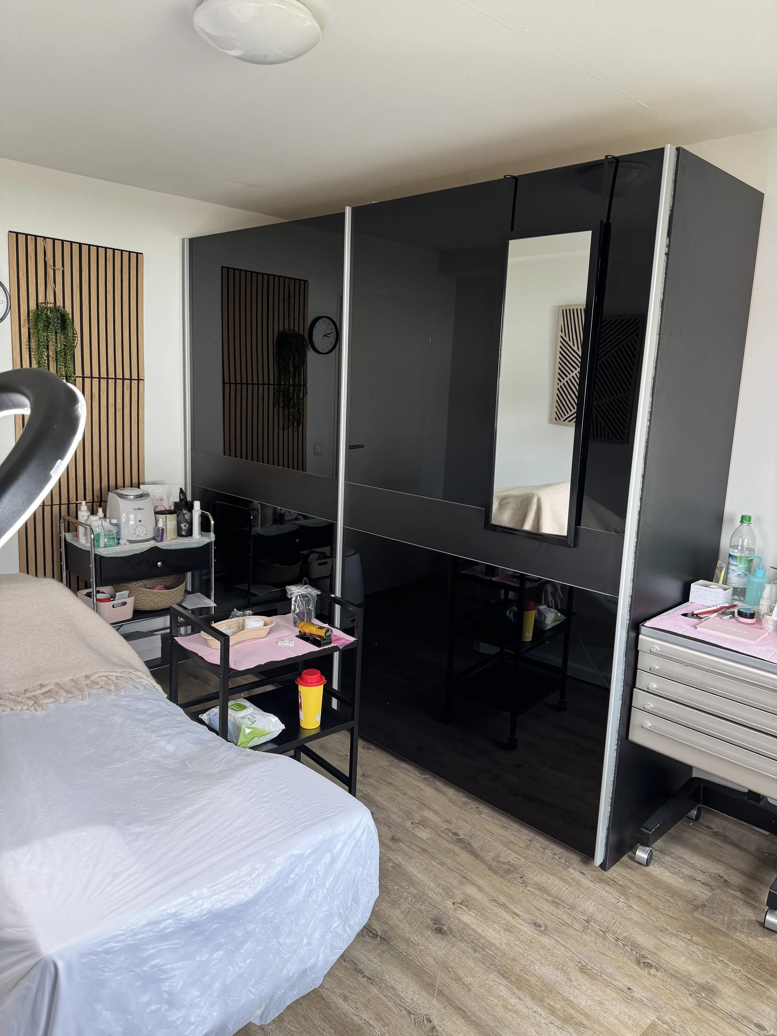 Ansicht eines Schlafzimmers mit einem frischen, weißen Laken auf dem Bett, einem schwarzen Nachttisch mit verschiedenen Gegenständen, einem Standspiegel, einem Trennwandsystem mit Spiegel, einem Pflanzenbild an der Wand, sowie einer kleinen Tischchen und einem Schrank mit Utensilien auf dem Boden.