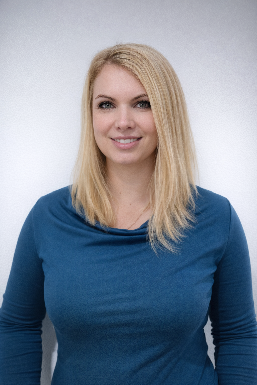 Porträt einer lachenden Frau mit blonden, geraden Haaren, trägt eine blaue Bluse, vor einem weißen Hintergrund.