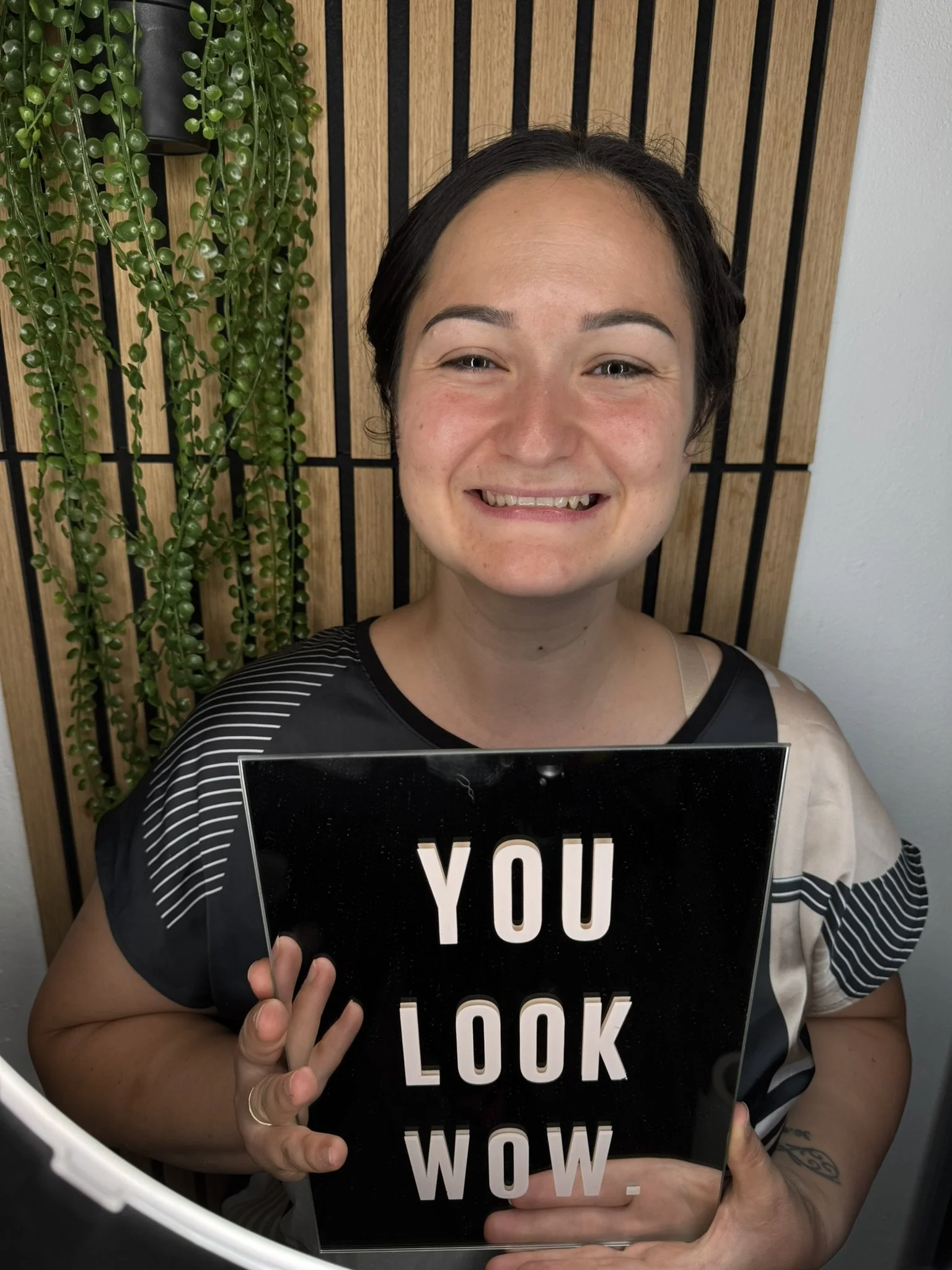 Eine lachende Frau hält eine schwarze Tafel mit der Aufschrift 'YOU LOOK WOW.' vor sich. Im Hintergrund sind grüne Pflanzen und eine Holzverkleidung sichtbar.