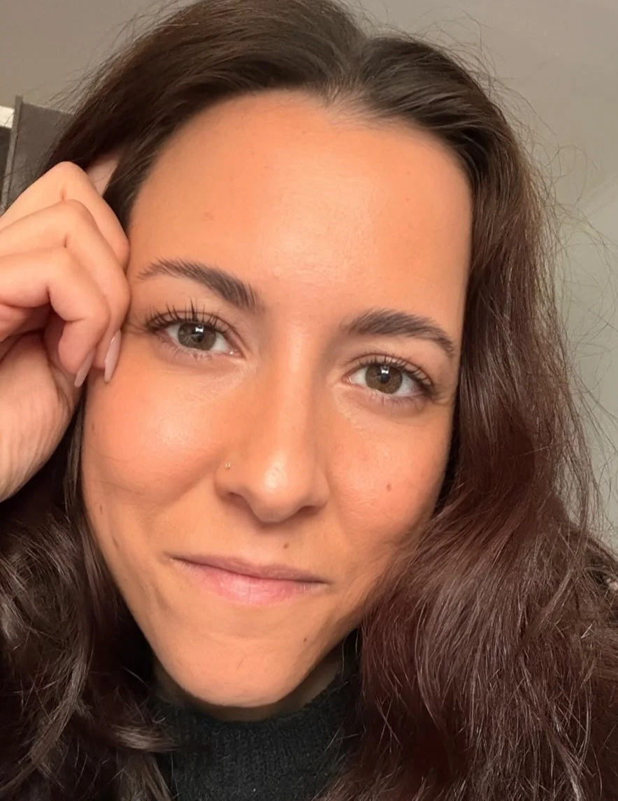 Nahaufnahme eines lächelnden Frauengesichts mit braunen, lockigen Haaren und natürlichem Make-up, die die Hand an die Seite ihres Gesichts legt.