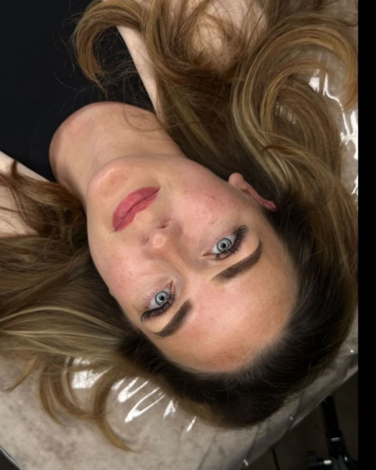 Eine Frau mit langen, lockigen, honigblonden Haaren und blauen Augen liegt auf einer glatten Oberfläche, blickt in die Kamera, trägt schwarzen Oberteil, natürliche Make-up, rote Lippen.