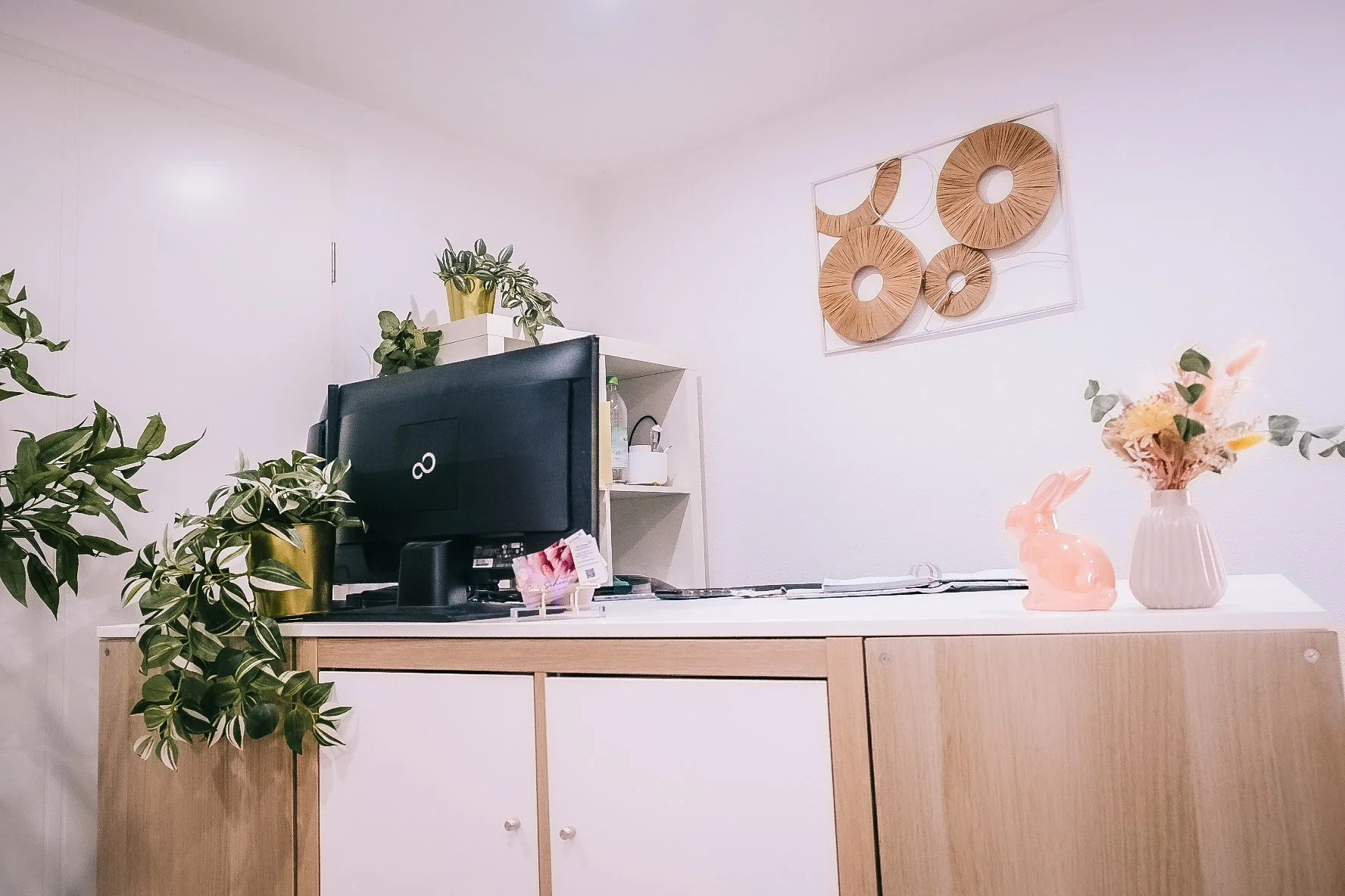 Küche mit weißen Wänden, einer Holzschrank vorne, Pflanzen in goldenen und weißen Töpfen, Wanddekor aus Holzringen, rosa Keramikhase und Vase mit Trockenblumen auf der Arbeitsfläche, Wasserkocher und Küchenutensilien.