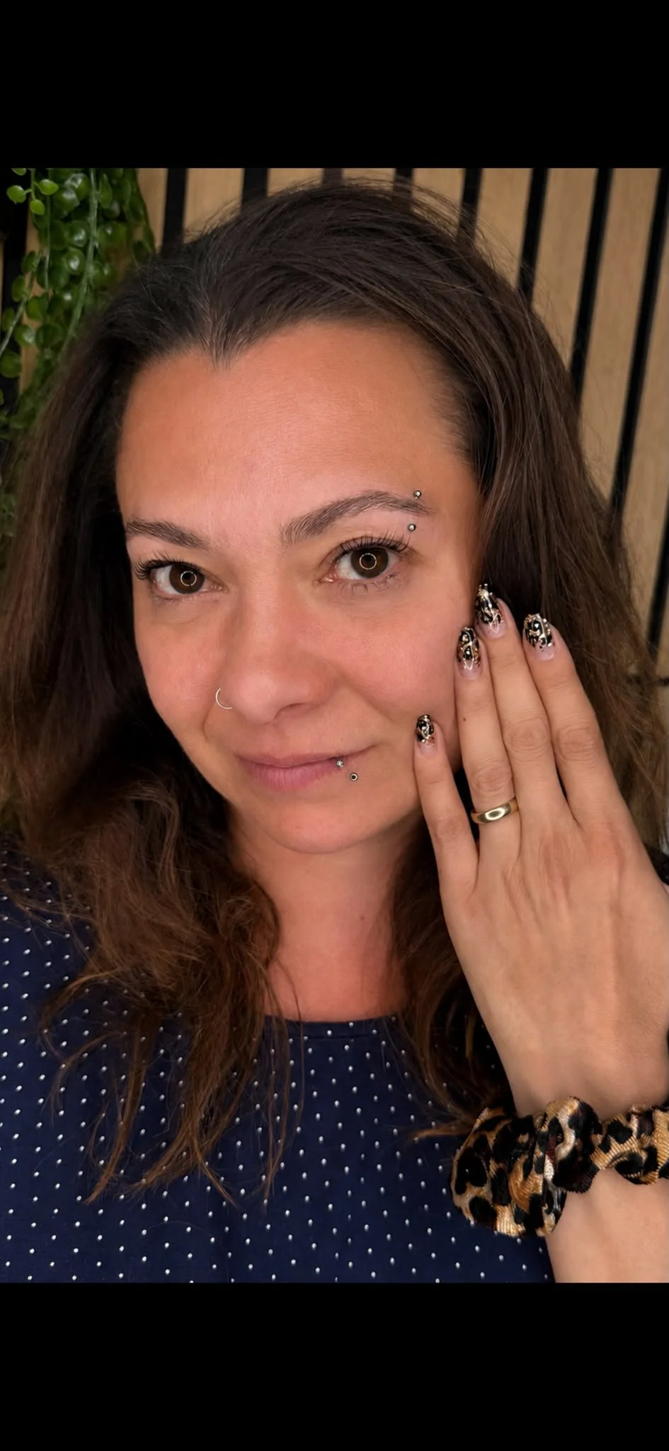 Nahaufnahme einer Frau, die ihre rechte Hand an die rechte Wange legt, mit schwarzer, weiß gepunkteter Bluse, langen braunen Haaren, Piercings im Gesicht und bunten Nagellack mit Muster, vor einem Holz- und Pflanzenhintergrund.