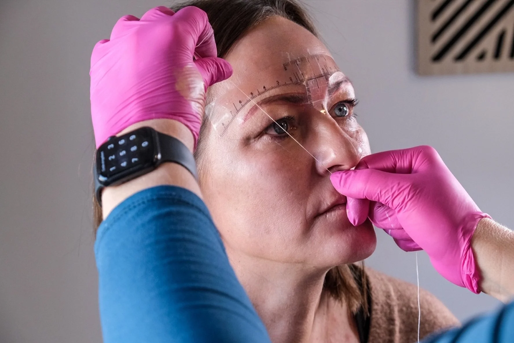Eine Frau wird für eine medizinische Untersuchung vorbereitet, trägt pinke Handschuhe, eine Uhr und hat Messgeräte auf der Stirn, während eine Person ihr die Nase berührt.