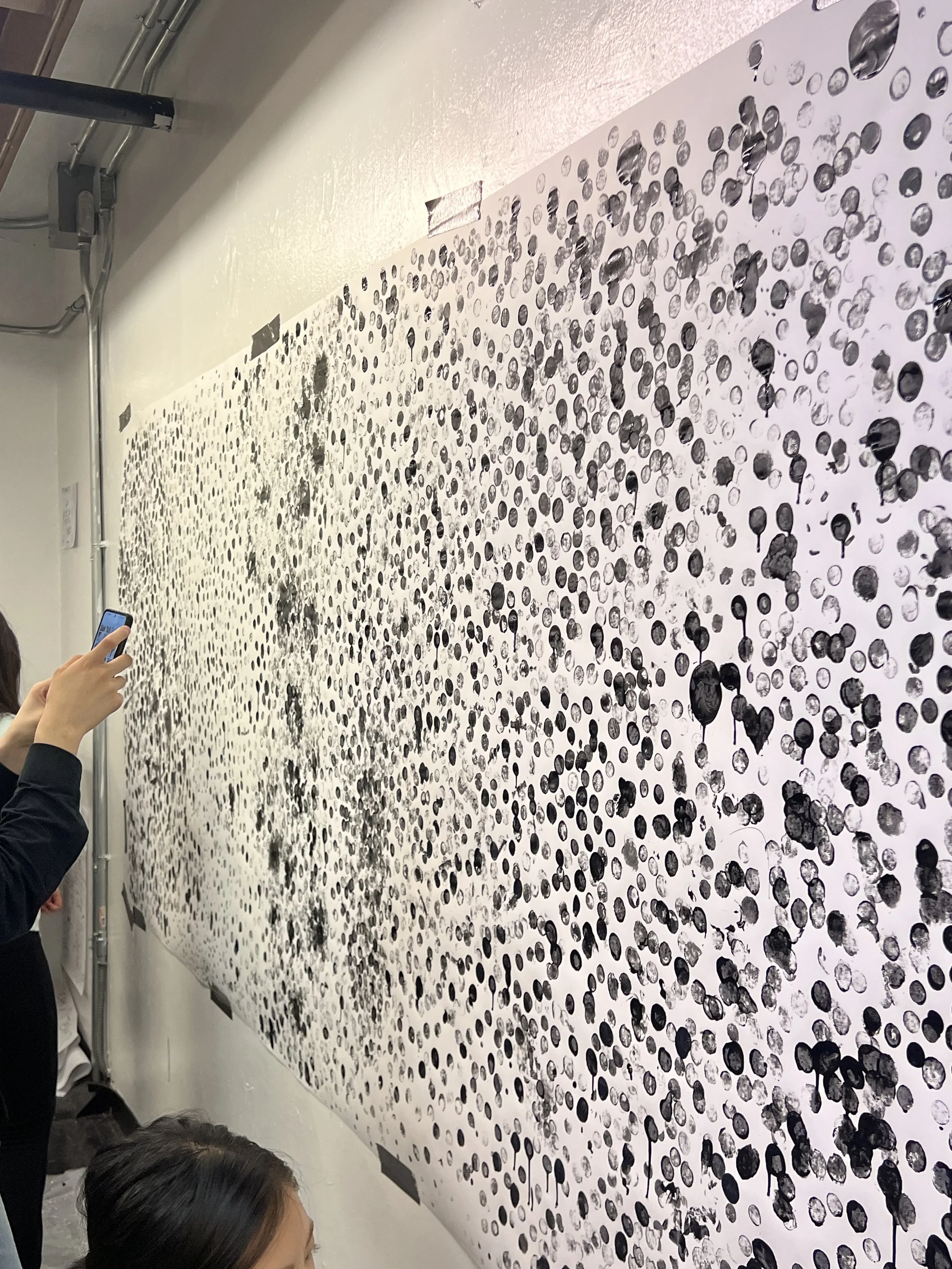 Large abstract black and white artwork with numerous black dots on white paper, taped to a white wall, with people viewing and photographing it.