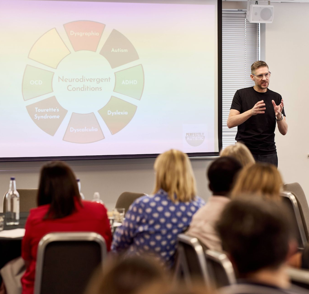 A man giving a presentation to an audience in a conference room, with a slide about neurodivergent conditions projected on the screen.