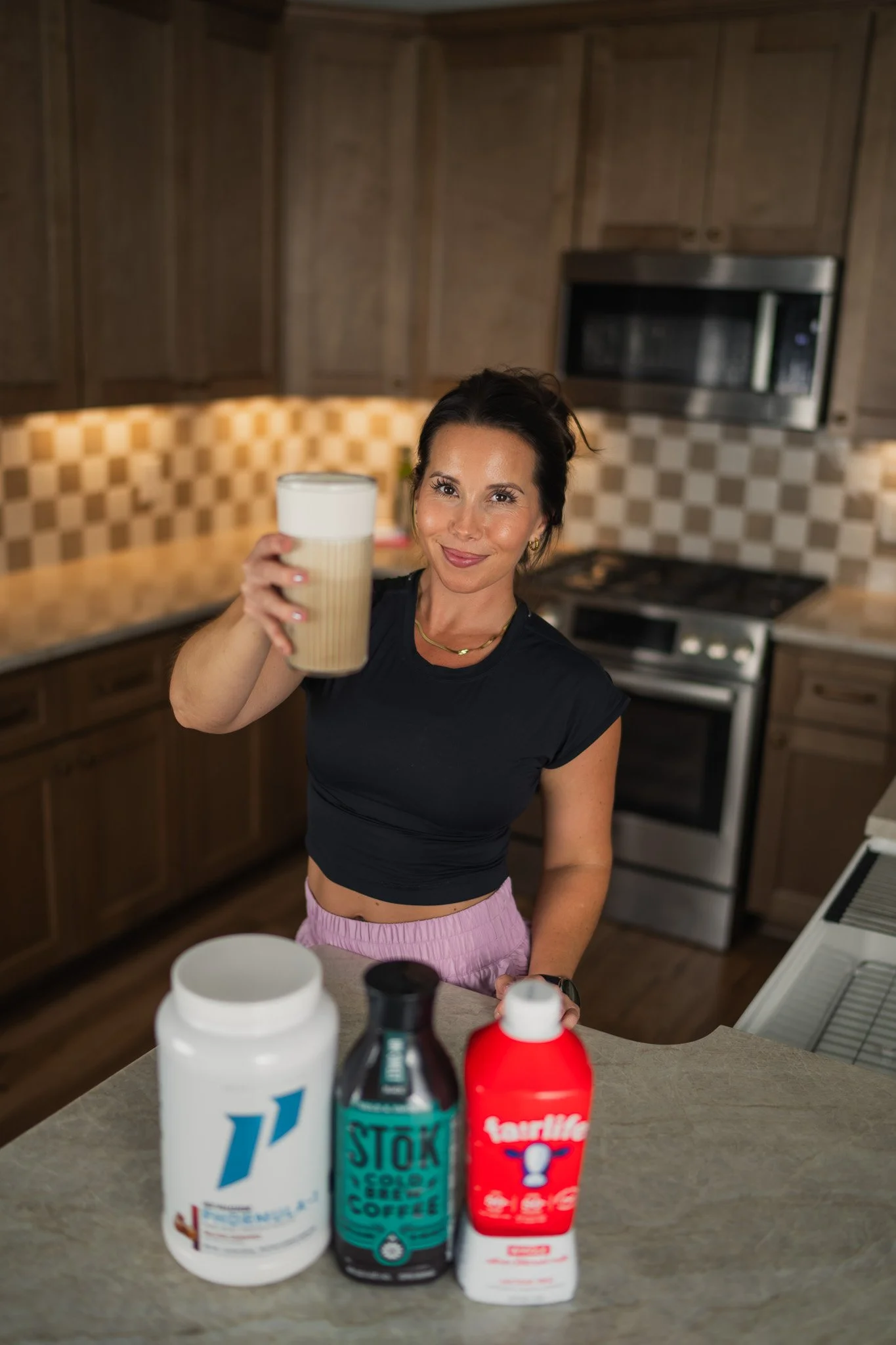 A woman in a black shirt and pink shorts holding up a beige cup in a kitchen with wooden cabinets and checkered backsplash, with various supplement bottles on the counter in front of her.
