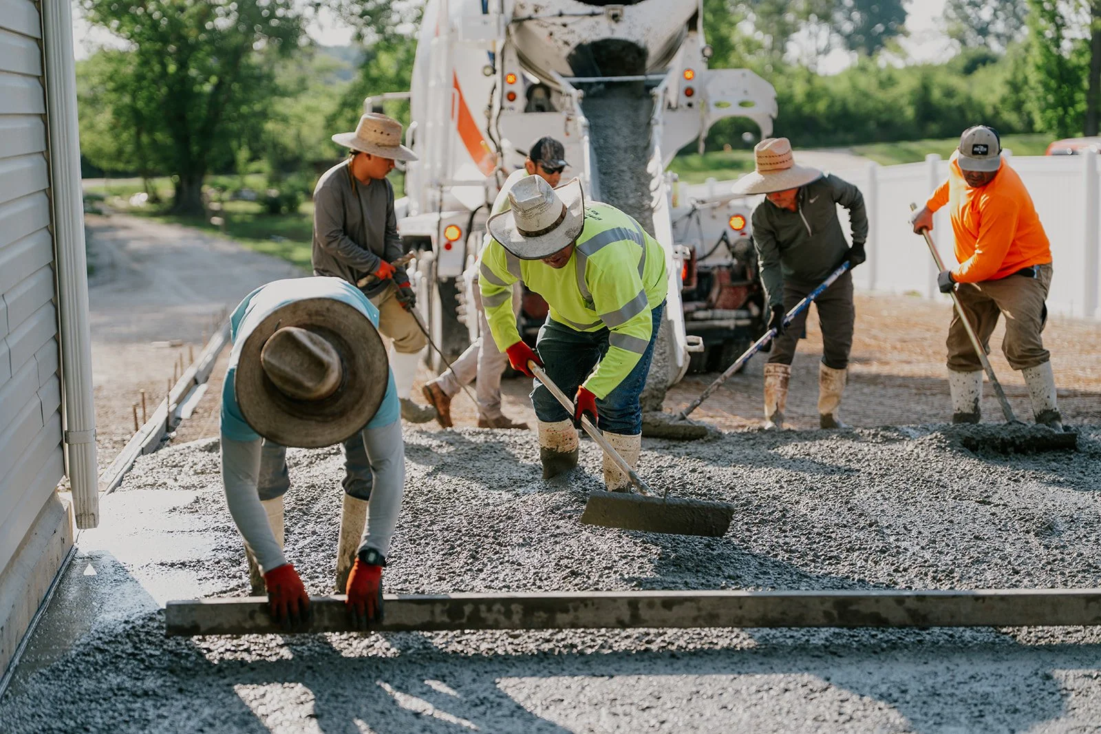 Construction workers pour and level concrete on a driveway, with a concrete mixer truck in the background and trees in the distance.