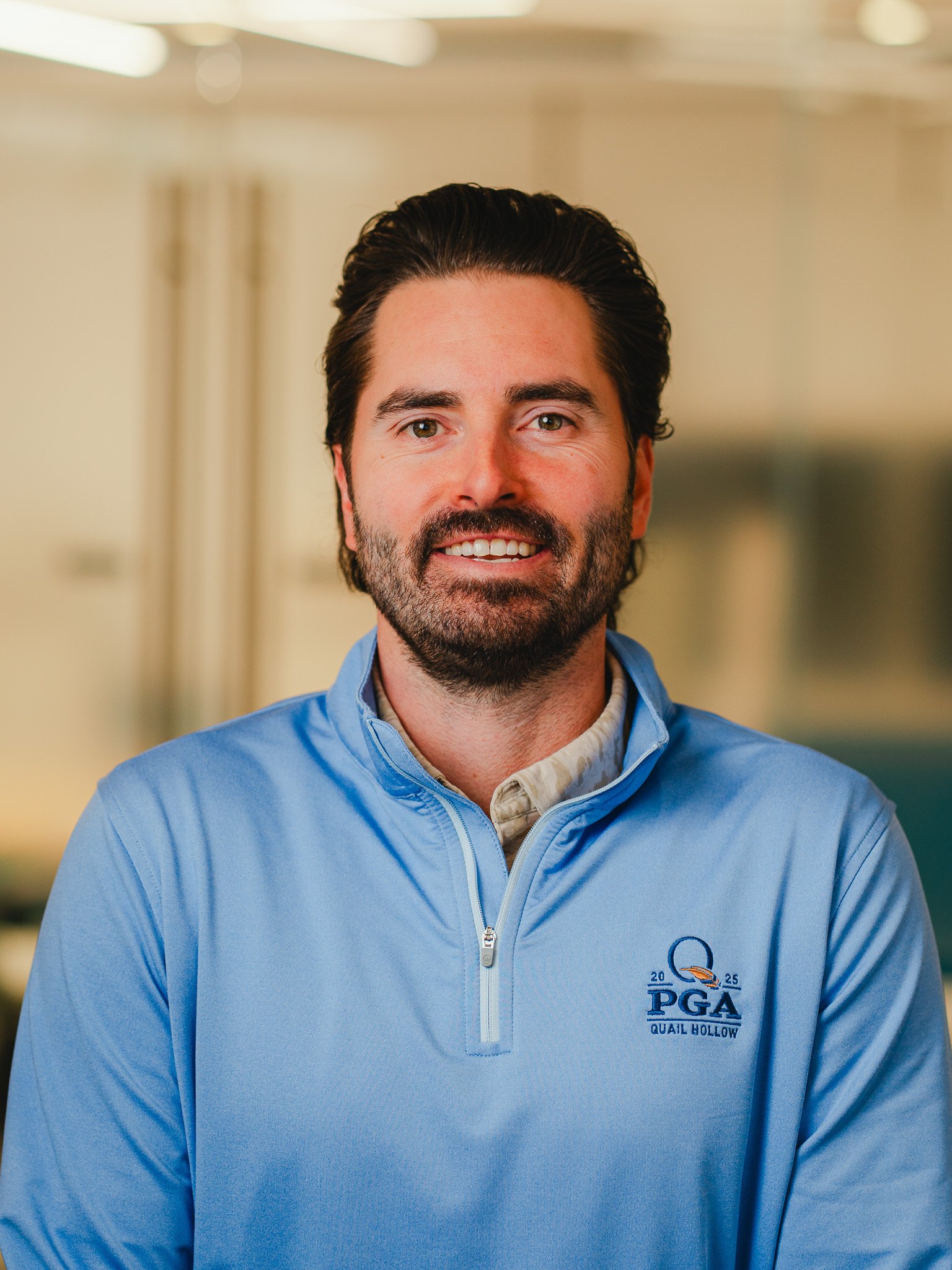 Close-up of a man with dark hair and beard wearing a light blue jacket with a PGA logo on it, smiling in an indoor setting.