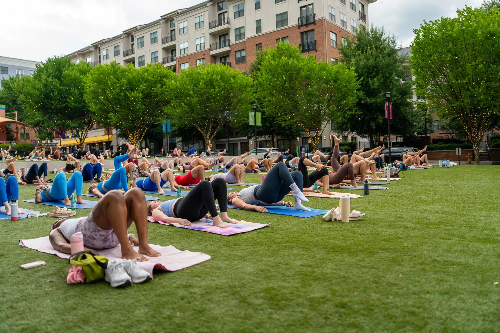 Many people practicing yoga outdoors on a grassy area with trees decorated with string lights, apartment buildings in the background, and cloudy sky.