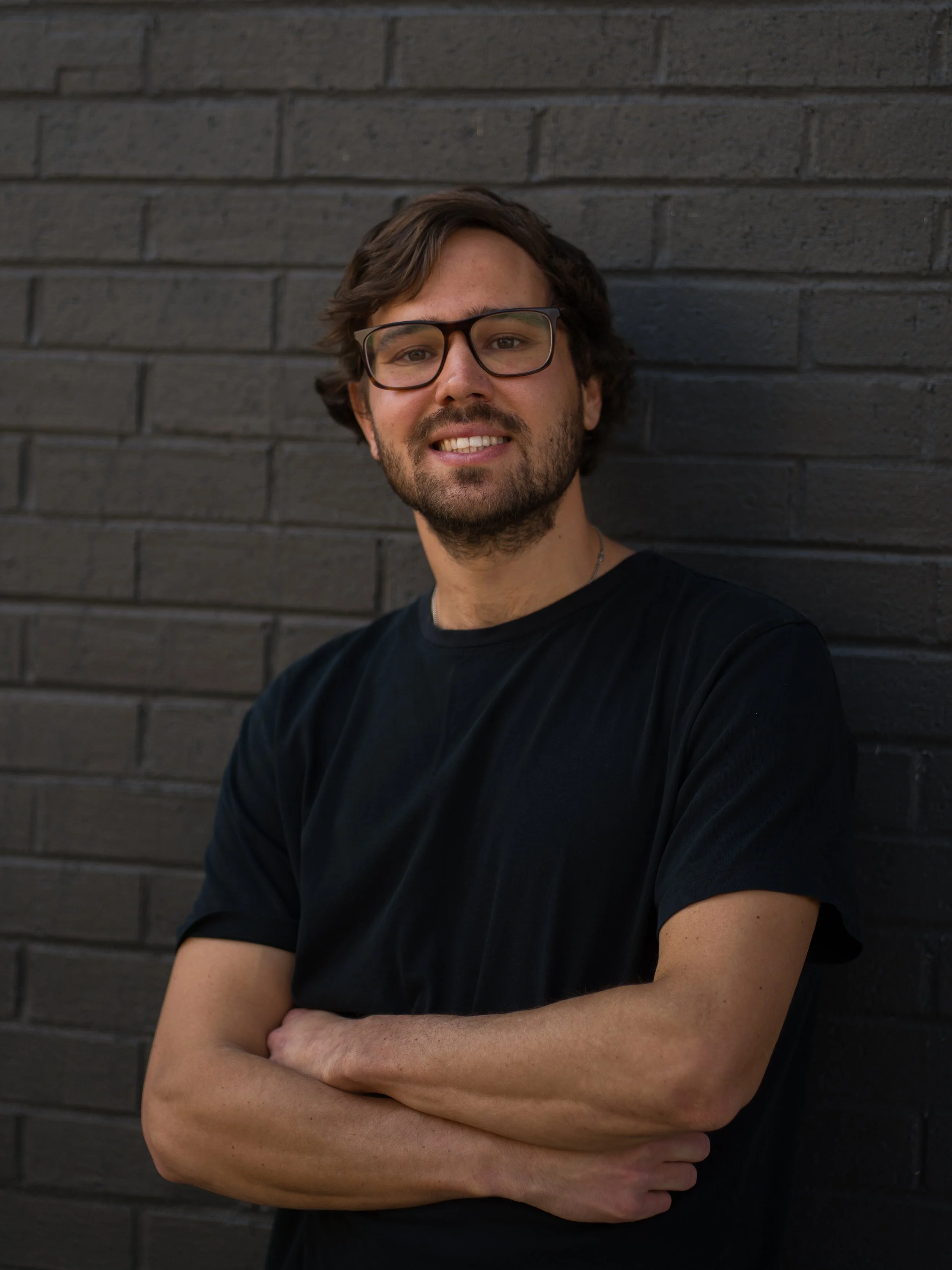 A man with glasses and a beard standing against a black brick wall, crossing his arms and smiling.