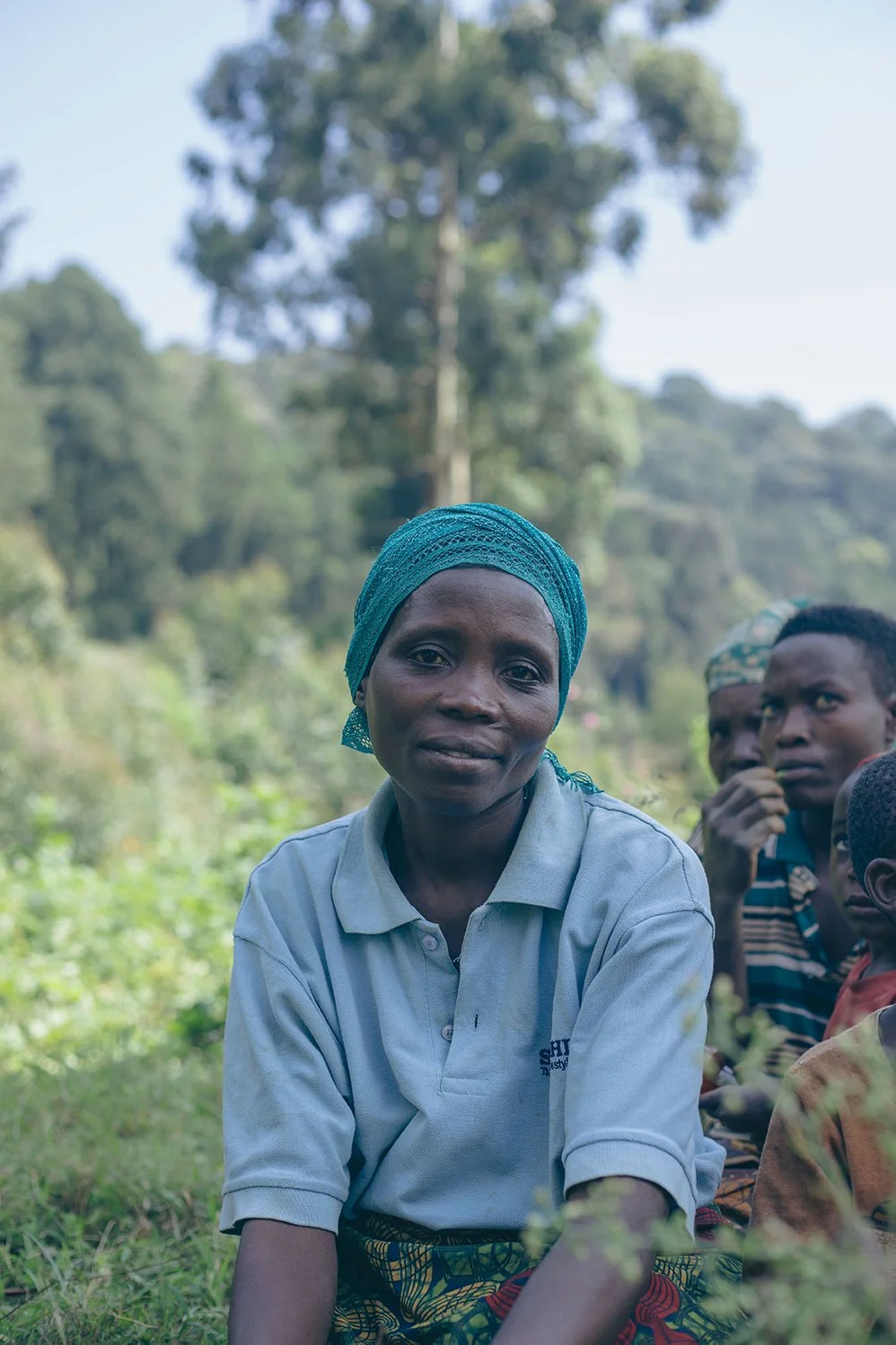 Femme burundaise assise dans un environnement naturel, portant un turban bleu et une chemise blanche.