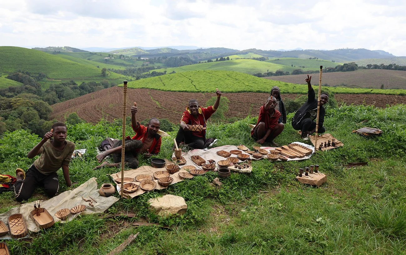 Groupe de personnes africaines posant avec des objets artisanaux en bois sur une colline verdoyante, arrière-plan de collines agricoles