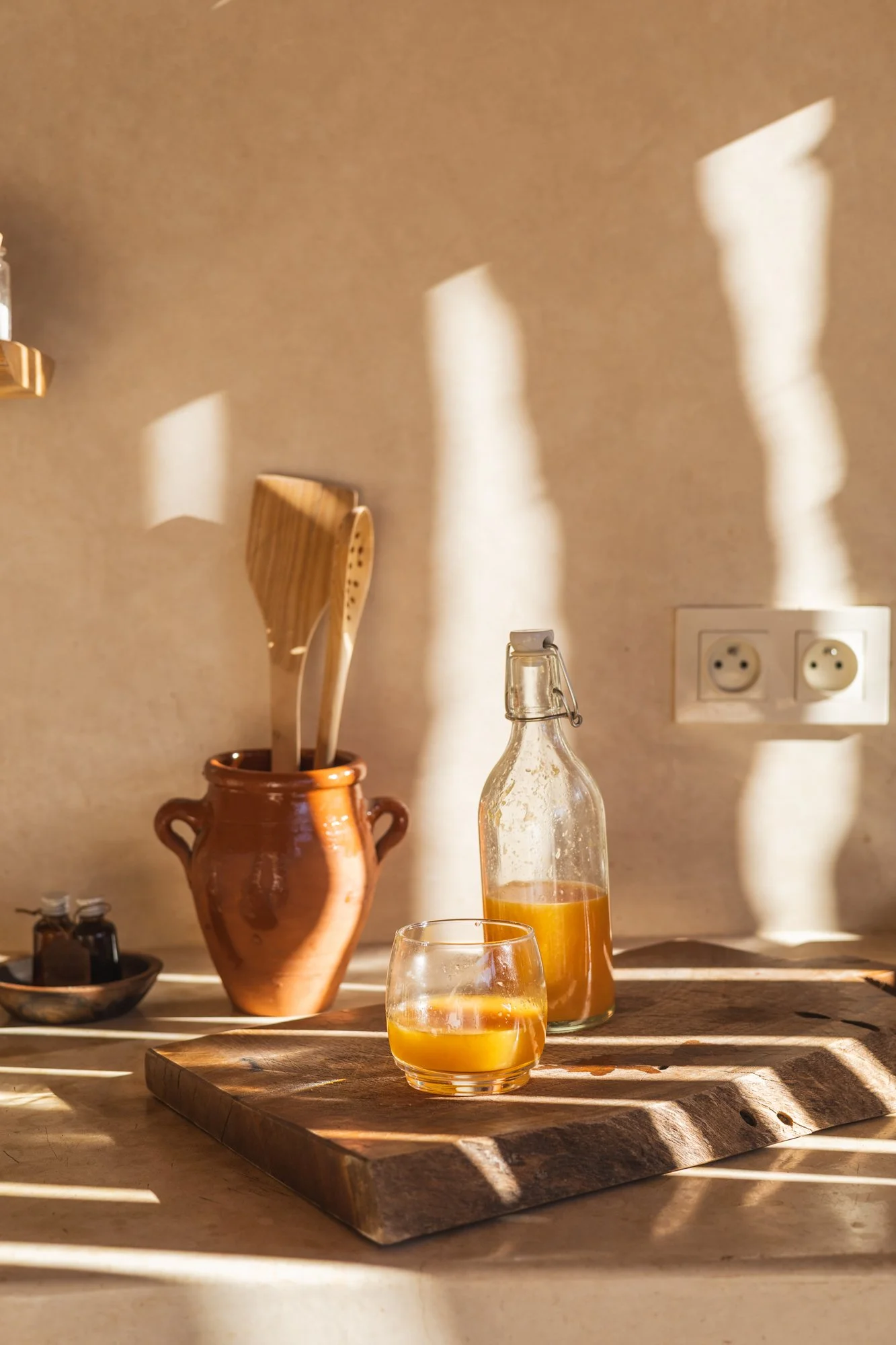 A glass bottle and a short glass filled with orange juice sit on a wooden cutting board on a kitchen countertop. Behind them is a clay pot with wooden kitchen utensils, and a beige wall with shadows cast by sunlight. There is a double power outlet on