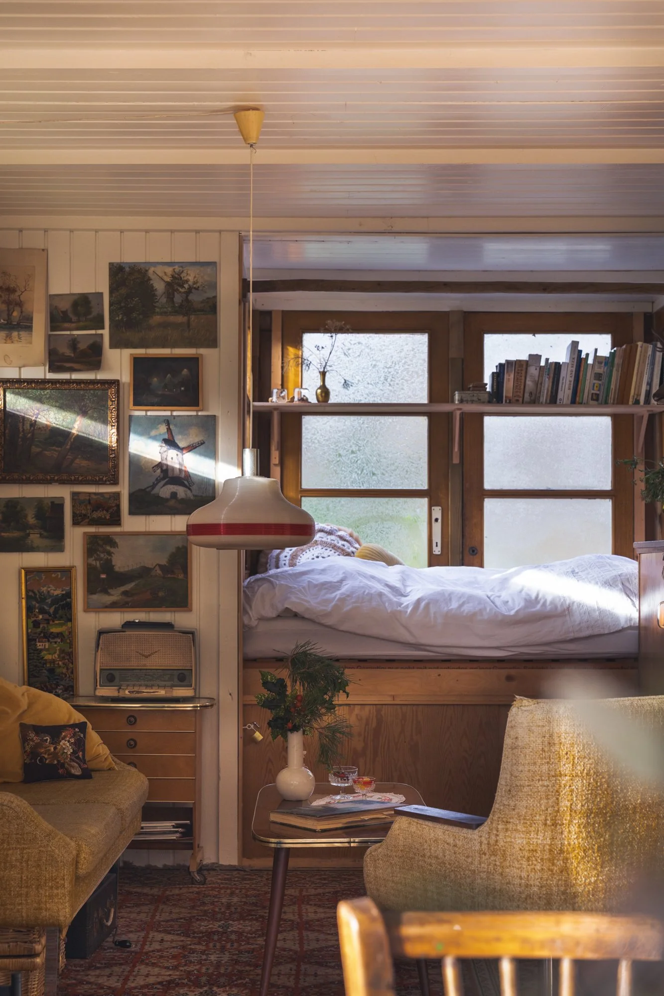 Cozy living room with a built-in bed by frosted windows, surrounded by vintage furniture, artwork on the walls, and a small table with a white vase and plants.