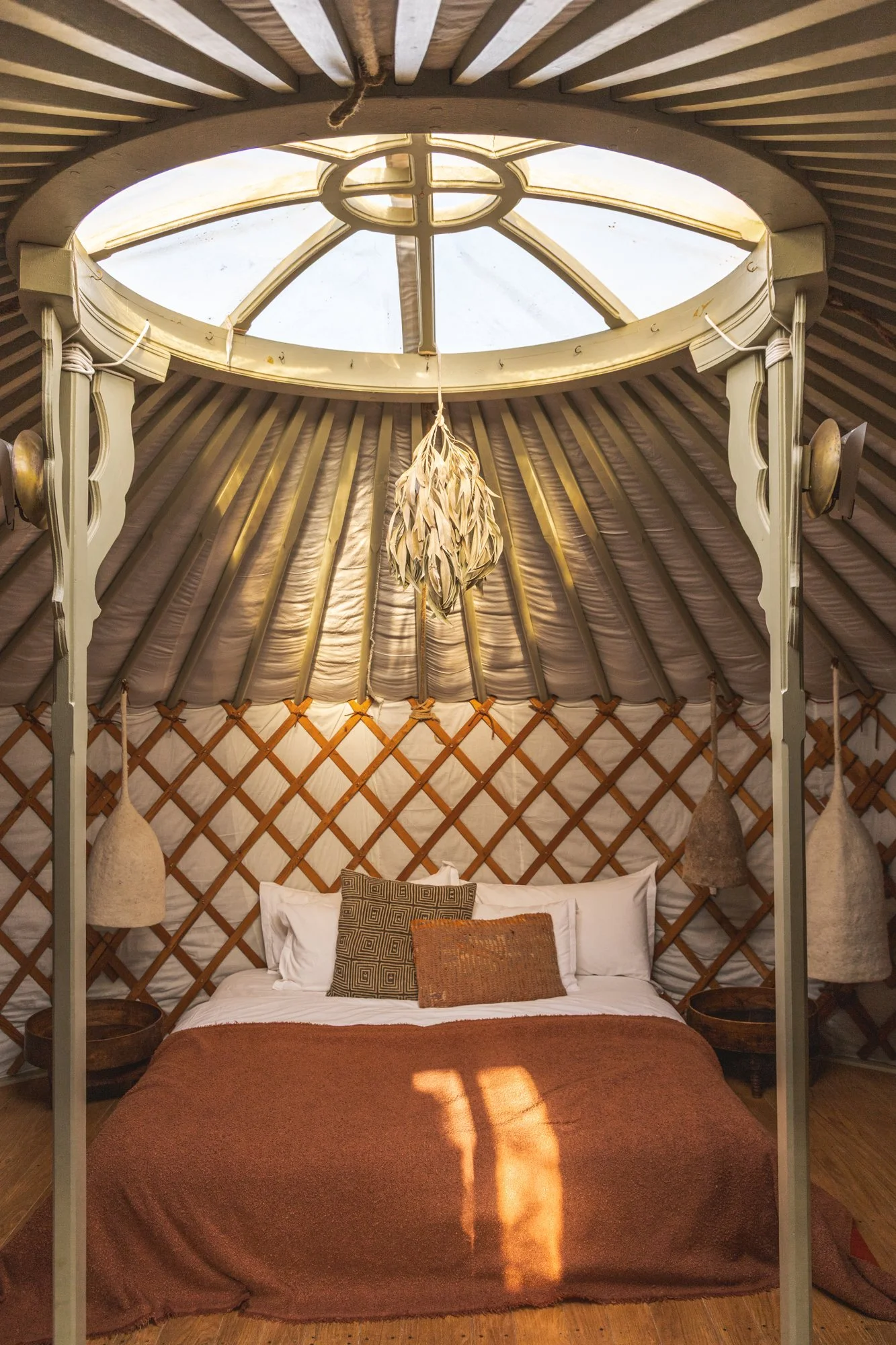 Interior of a yurt with a bed and rustic decor, including hanging lamps and pillows, under a circular skylight.