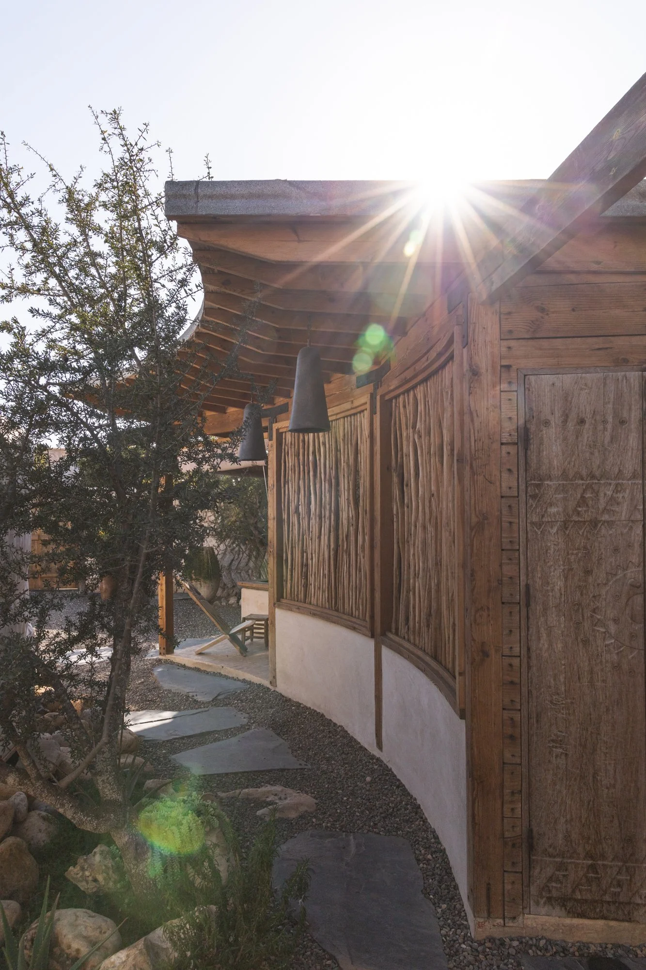 Sunlight shining on a wooden exterior wall of a building with a curved design, a small tree on the left, stone pathway on the ground, and a clear sky above.