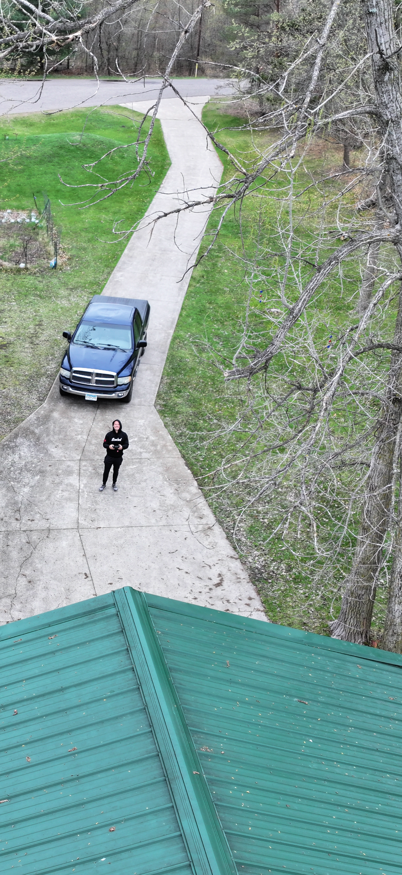 A person standing on a concrete driveway with a parked blue pickup truck nearby, a green roof in the foreground, and leafless trees along a grassy area adjacent to a road.