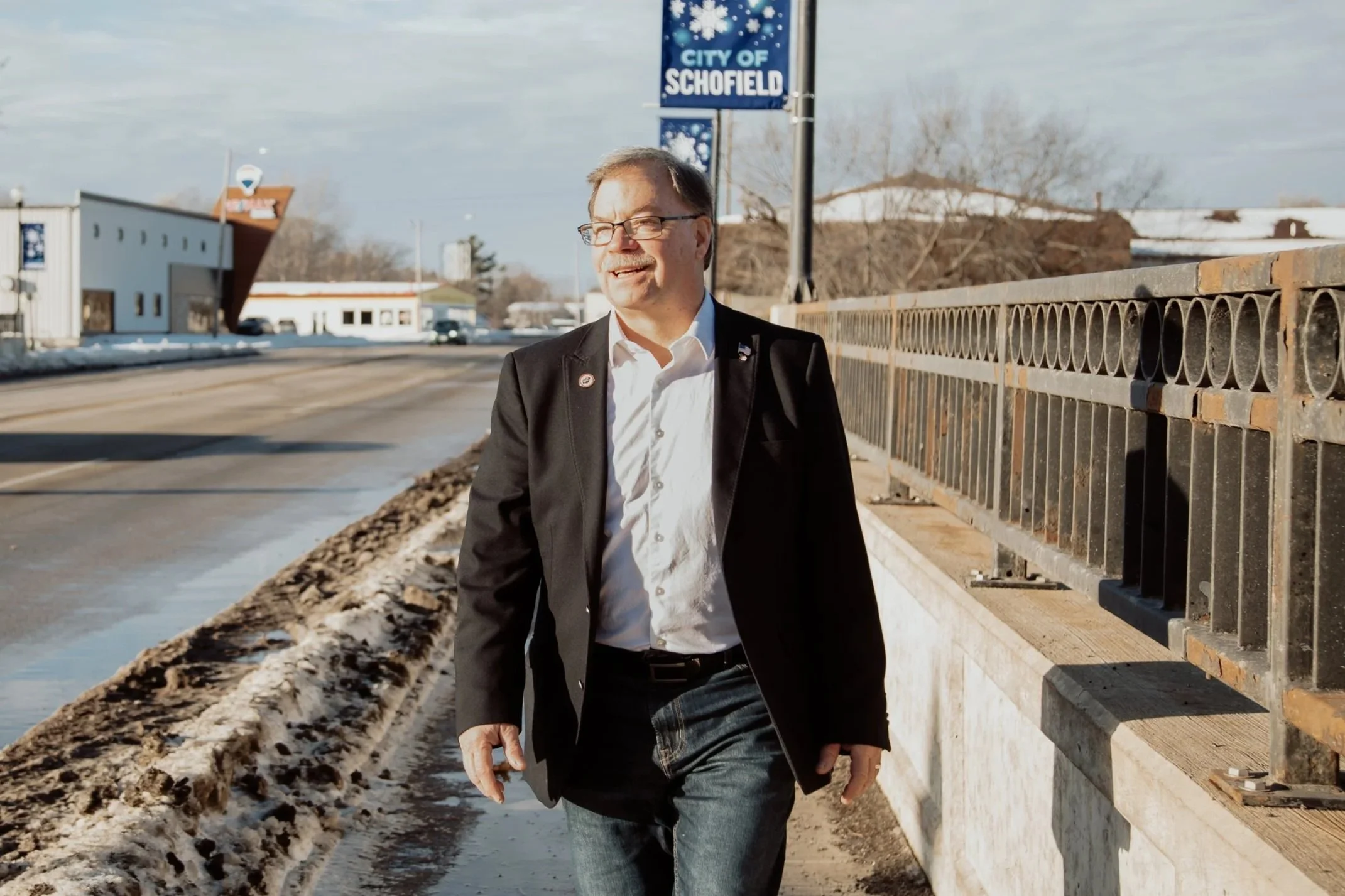 A man in a suit jacket and glasses walking along a sidewalk with a snow bank on one side and a black metal fence on the other, under a sign that reads 'City of Schofield' with snowflake graphics.
