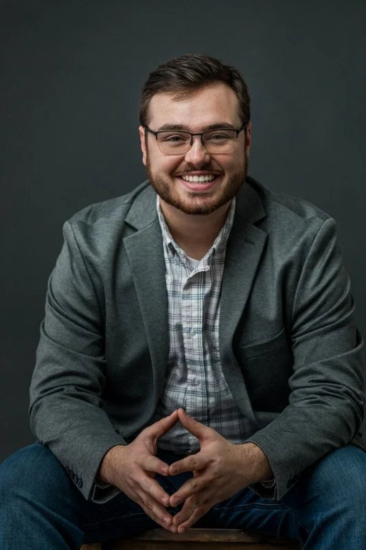 Portrait of a smiling man with glasses, a beard, in a gray blazer and checkered shirt, sitting against a dark background.