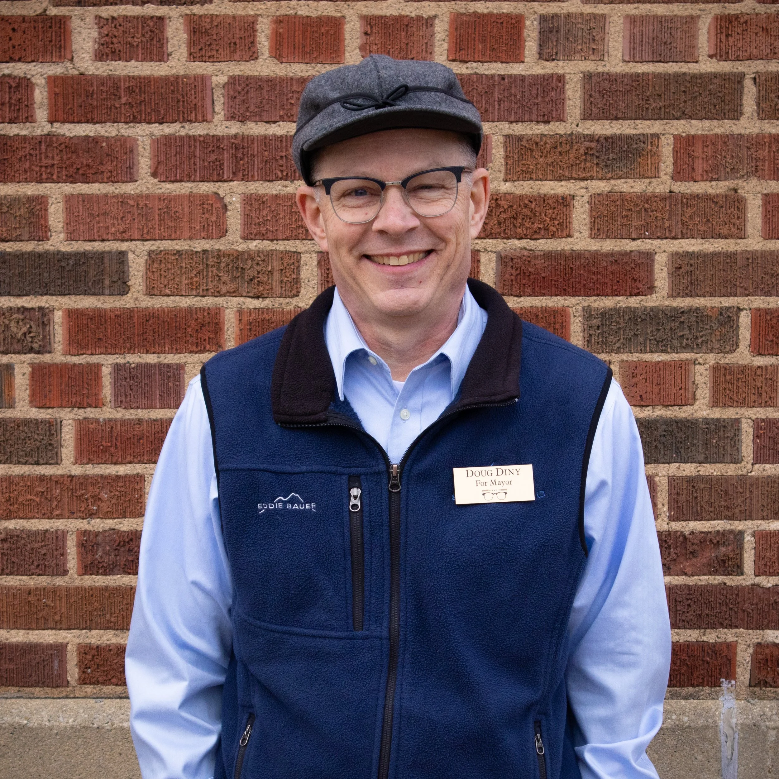 Wausau Mayor Doug Diny smiling in front of a brick wall wearing glasses, a gray cap, blue vest, light blue shirt, and a name tag.
