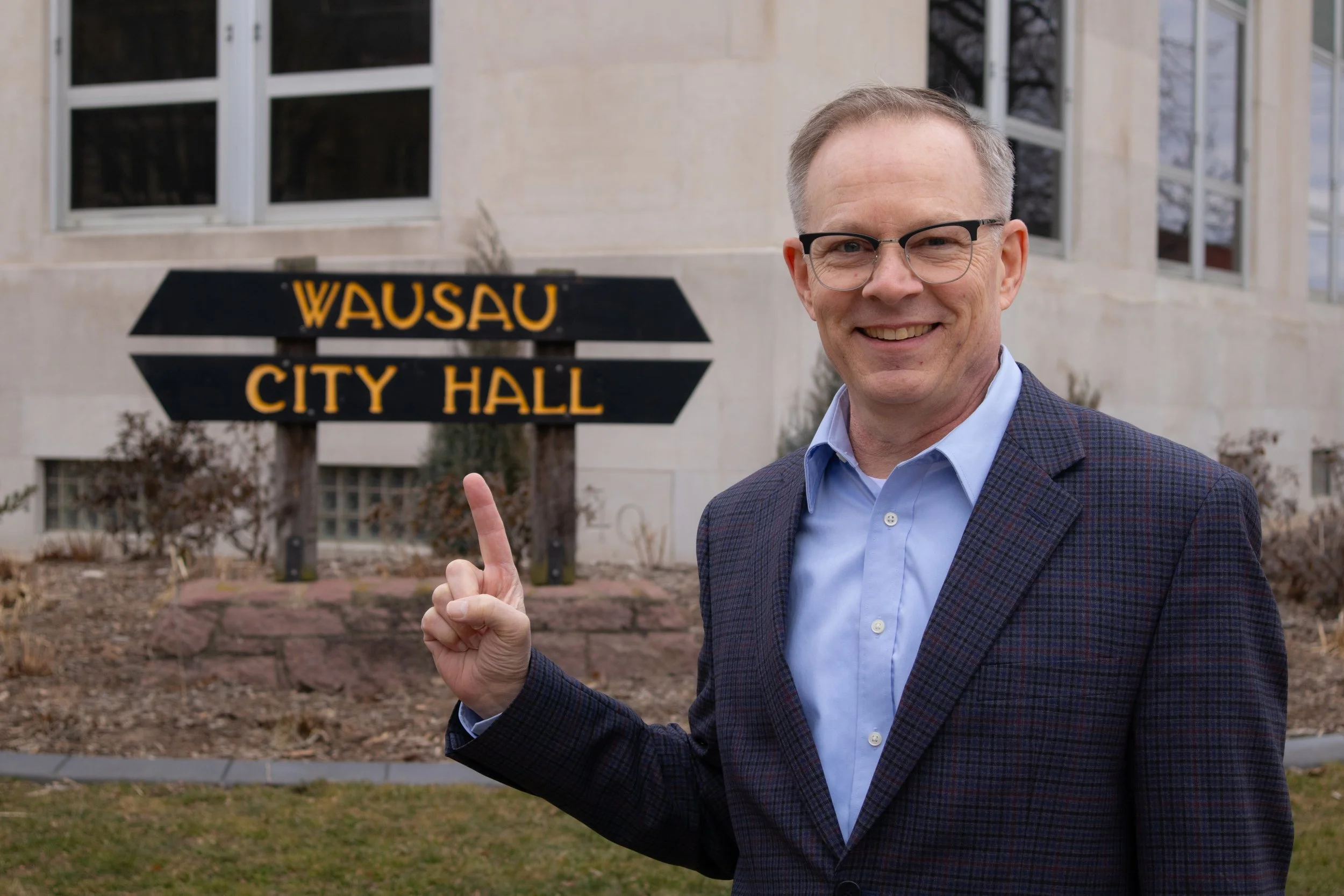 A man with glasses and a checkered blazer smiling and pointing at a sign that reads 'WAUSAU CITY HALL' outside a building with large windows.