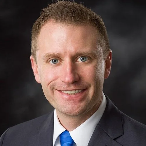 Portrait of a man with short light brown hair, blue eyes, wearing a white shirt, blue tie, and a dark suit, smiling against a dark background.