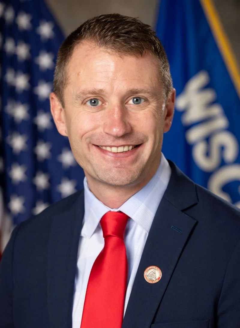 A smiling man in a suit with a red tie, standing in front of American flags and another blue flag.