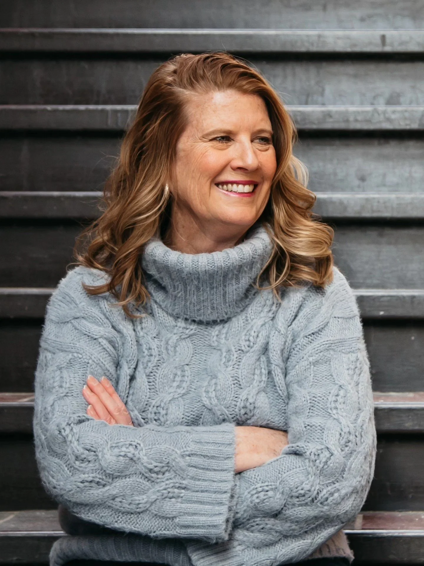 A middle-aged woman with wavy, shoulder-length reddish-brown hair, wearing a cozy gray cable-knit turtleneck sweater, standing on dark wooden stairs with a broad smile, arms crossed.