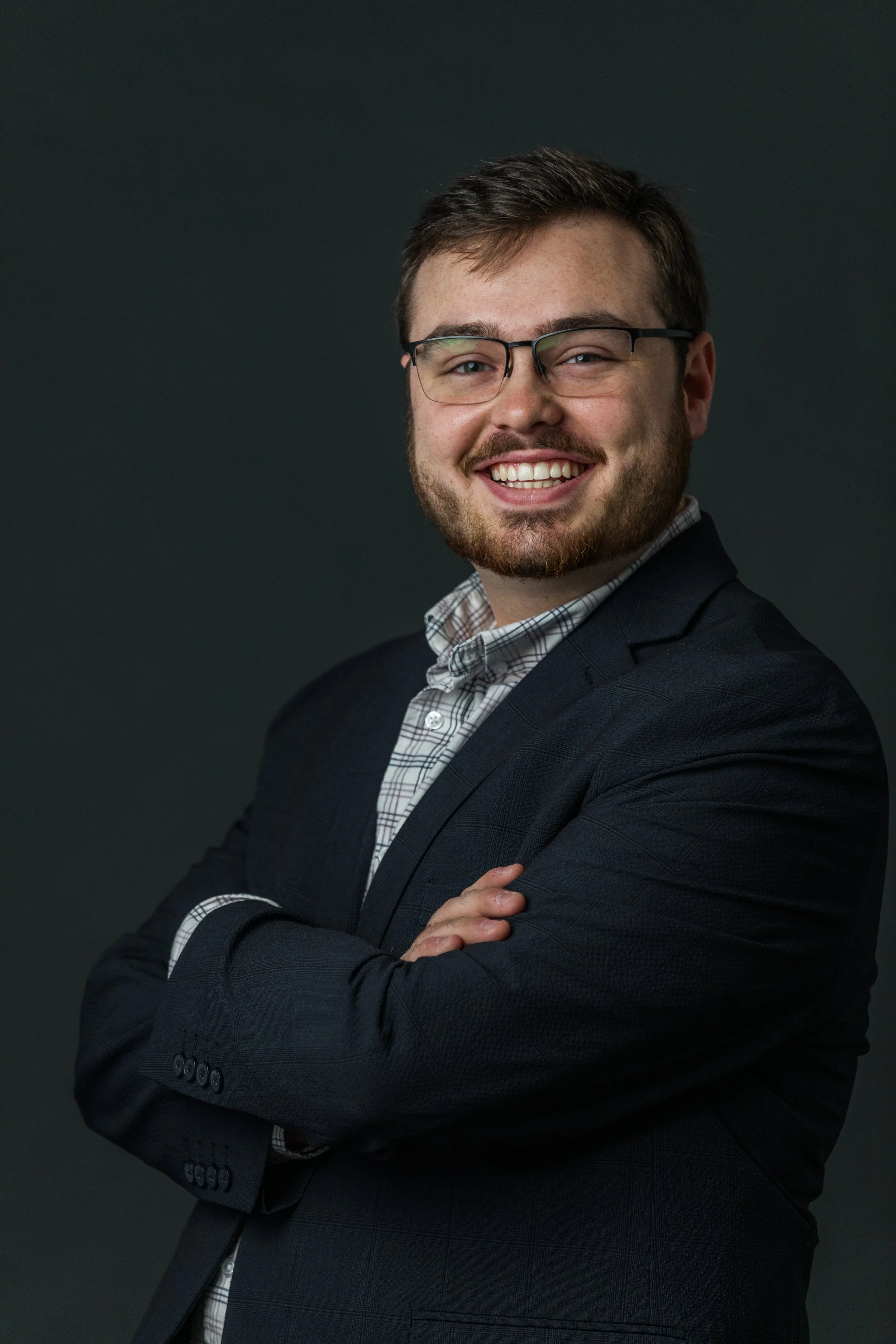 A man in a dark suit and plaid shirt smiling with his arms crossed against a dark background.