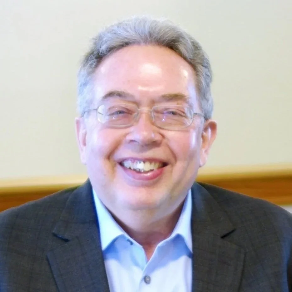 Smiling middle-aged man wearing glasses, a blue shirt, and a dark blazer in an indoor setting.
