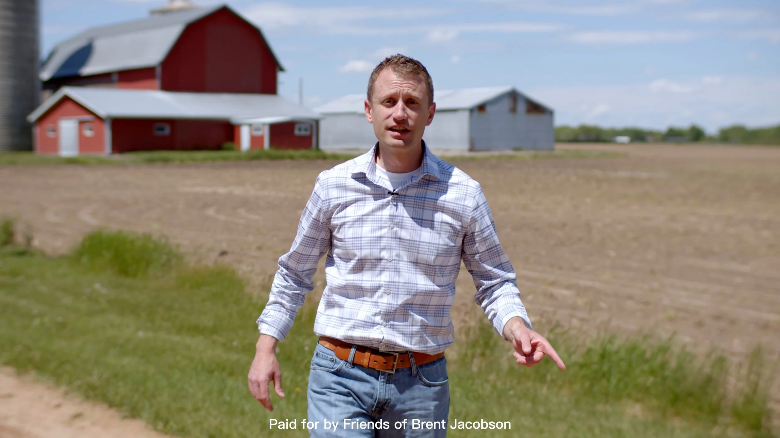 A man in a plaid shirt walking outdoors on a farm with red and gray barns in the background.