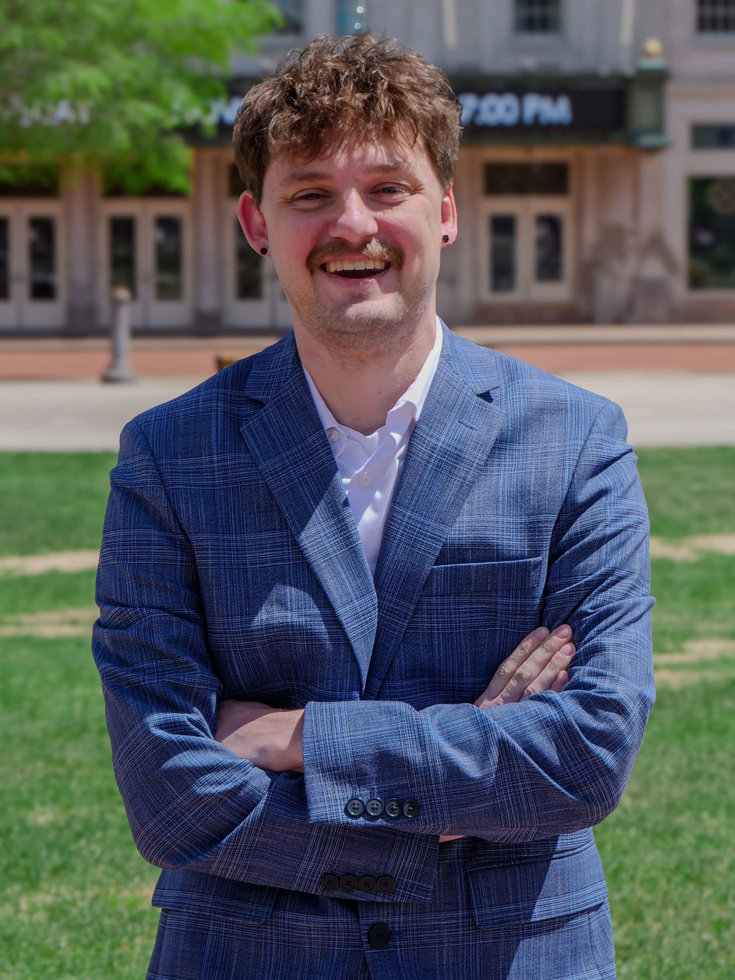 A young man with curly brown hair, wearing a blue checkered suit and white shirt, smiling with arms crossed outdoors near a grassy area and a building in the background.